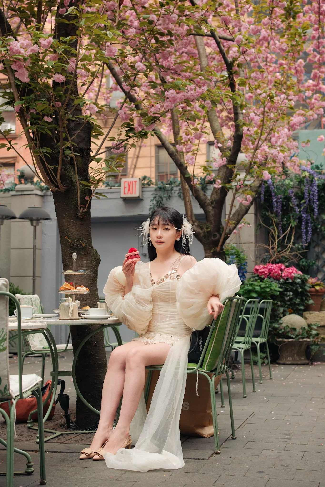 A young woman dressed in a white, vintage-style dress with puffy sleeves and a sheer train sitting at a garden table. She is holding a pink dessert, possibly a cupcake, and appears to be looking at the camera. The scene is set outdoors in a garden wi