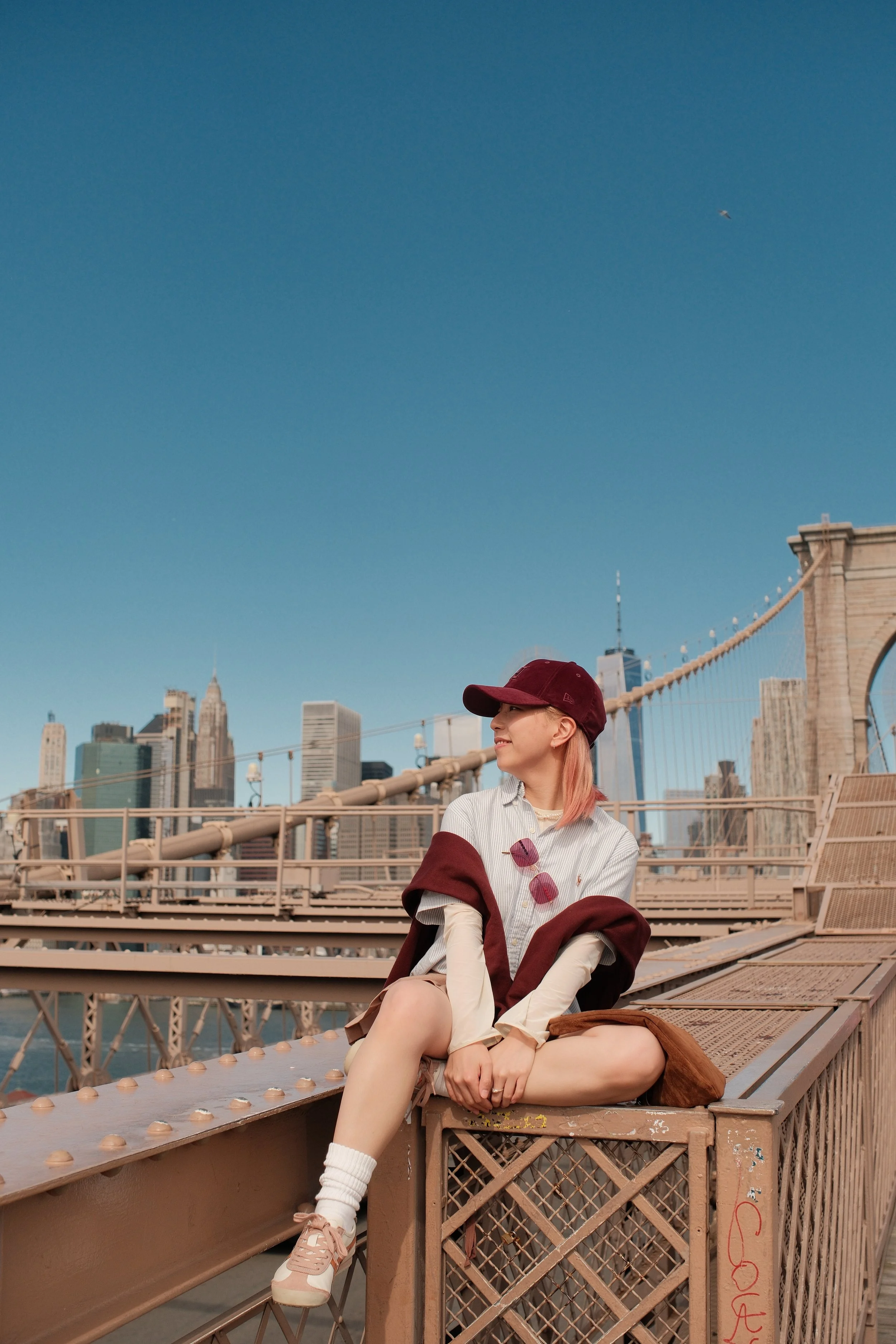 A young woman sitting on a metal structure on the Brooklyn Bridge in New York City, with the Manhattan skyline in the background, wearing a white striped shirt, a maroon cap, glasses, and sneakers, smiling and looking to the side.
