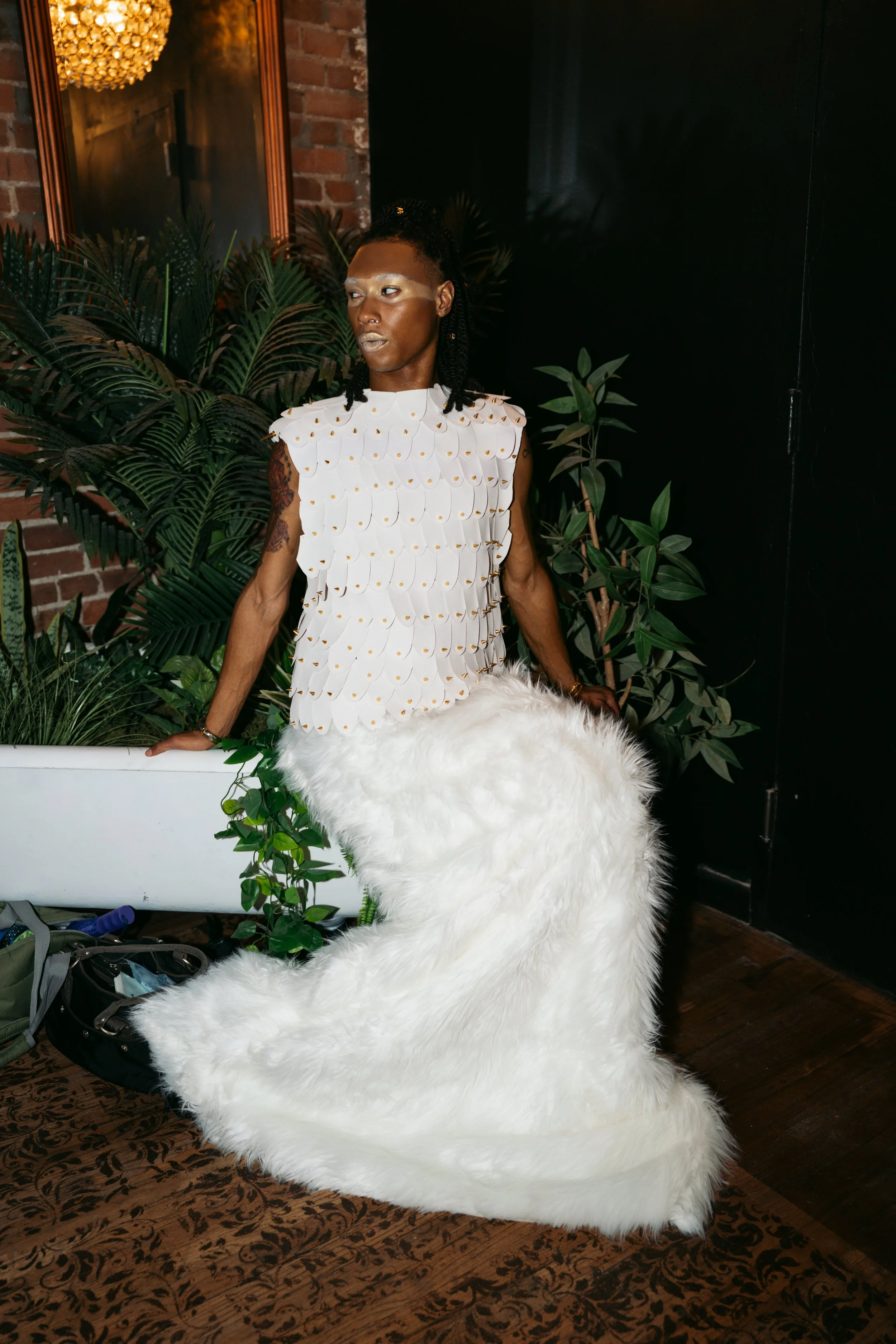 A person with dark skin and dreadlocks, wearing a white textured top with gold accents, standing indoors next to a large white fluffy rug and plants, with a brick wall and black wall in the background.