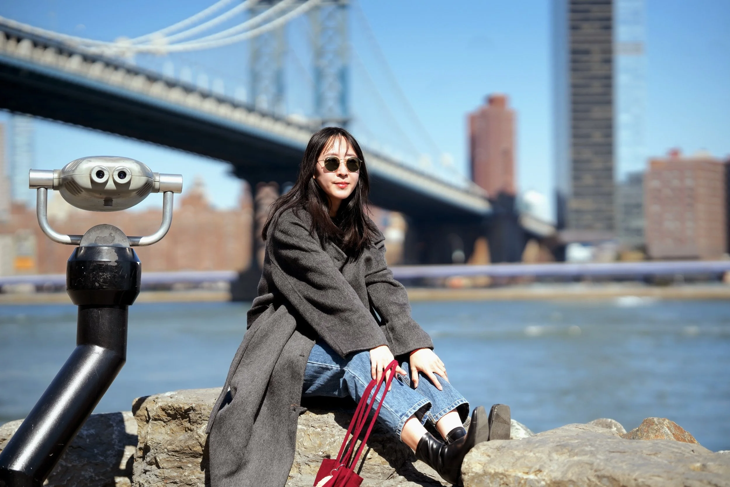 A woman sitting on rocks by the water with a bridge in the background, wearing sunglasses, a gray coat, and jeans, with a coin-operated binocular viewer nearby.