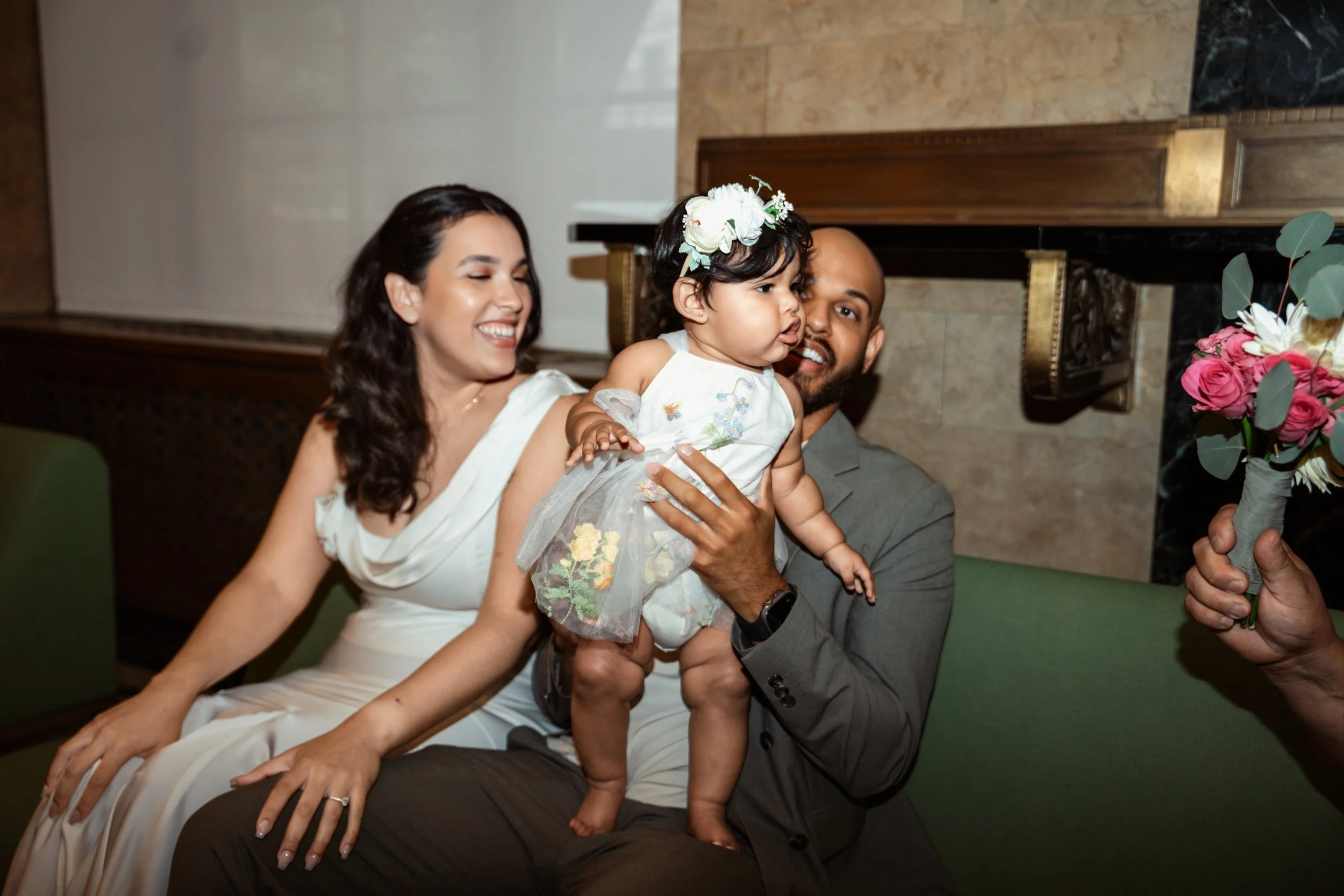 A woman in a white dress, a man in a gray suit, and a young girl wearing a flower crown are smiling and celebrating together indoors. The woman is seated, the man is holding the girl, and they are all happy.