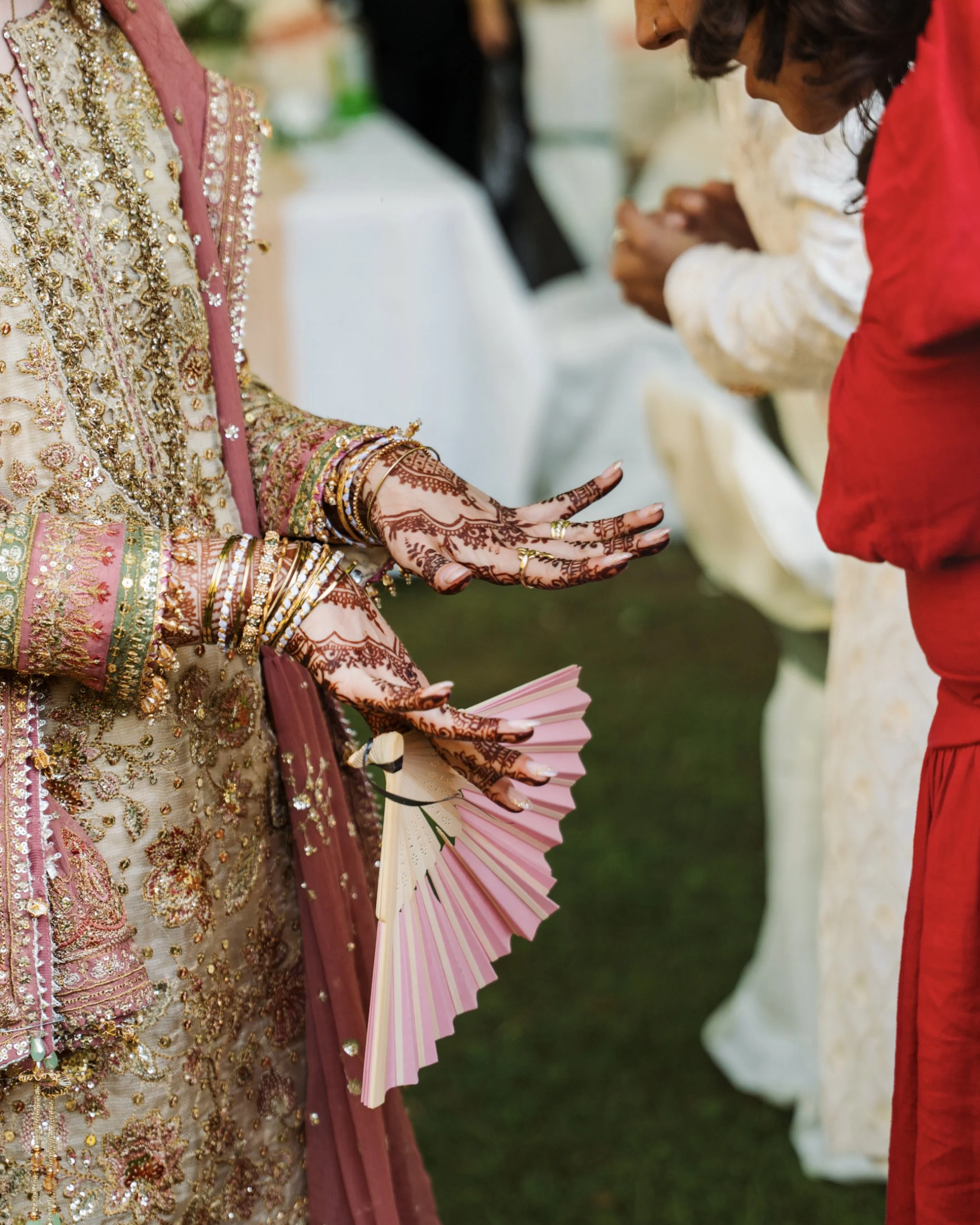Close-up of a woman dressed in traditional South Asian bridal attire, showing intricate gold embroidery and jewelry, holding a pink and cream paper fan, with another person in red and beige attire seen in the background.