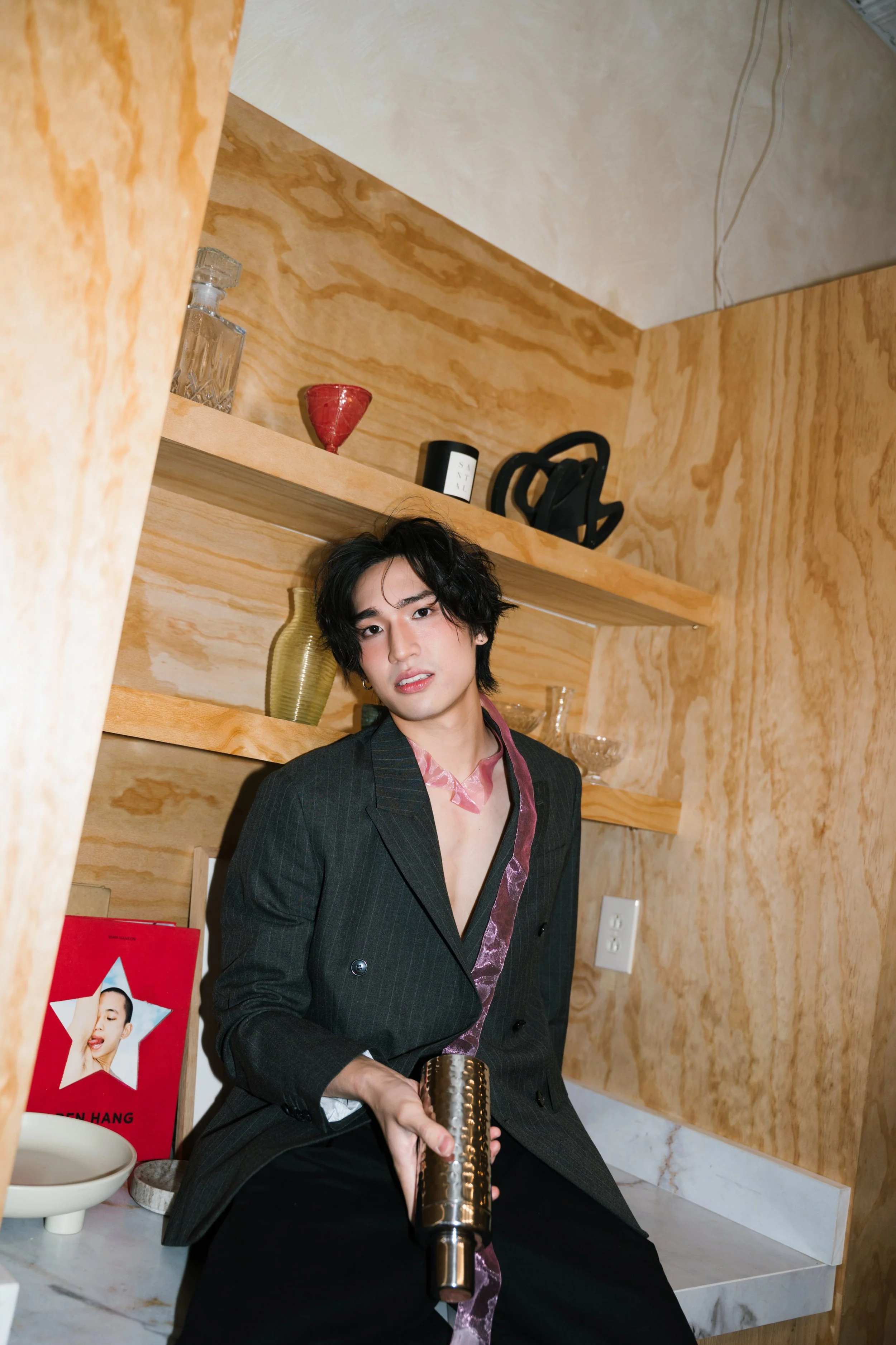 A young man with black, wavy hair sits on a marble counter in a room with wood-paneled walls. He wears a dark pinstripe blazer and a pink, patterned scarf. He holds a metallic object in his hand, and there are decorative items and a poster of a woman