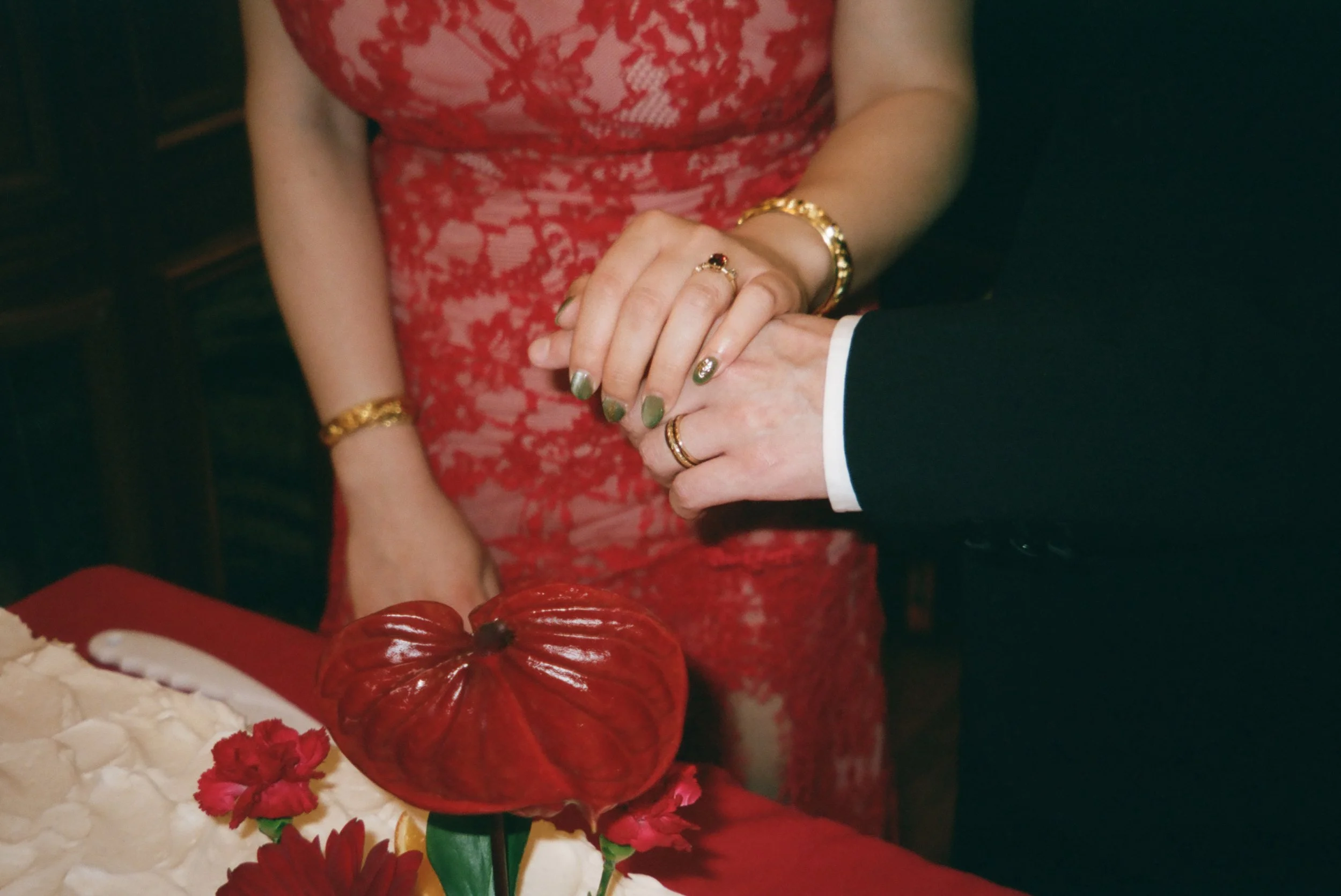 Close-up of a woman in a red lace dress holding hands with a man in a dark suit; they are wearing gold jewelry and rings. The scene appears to be a wedding or formal event, with table decorations and floral arrangements in the foreground.