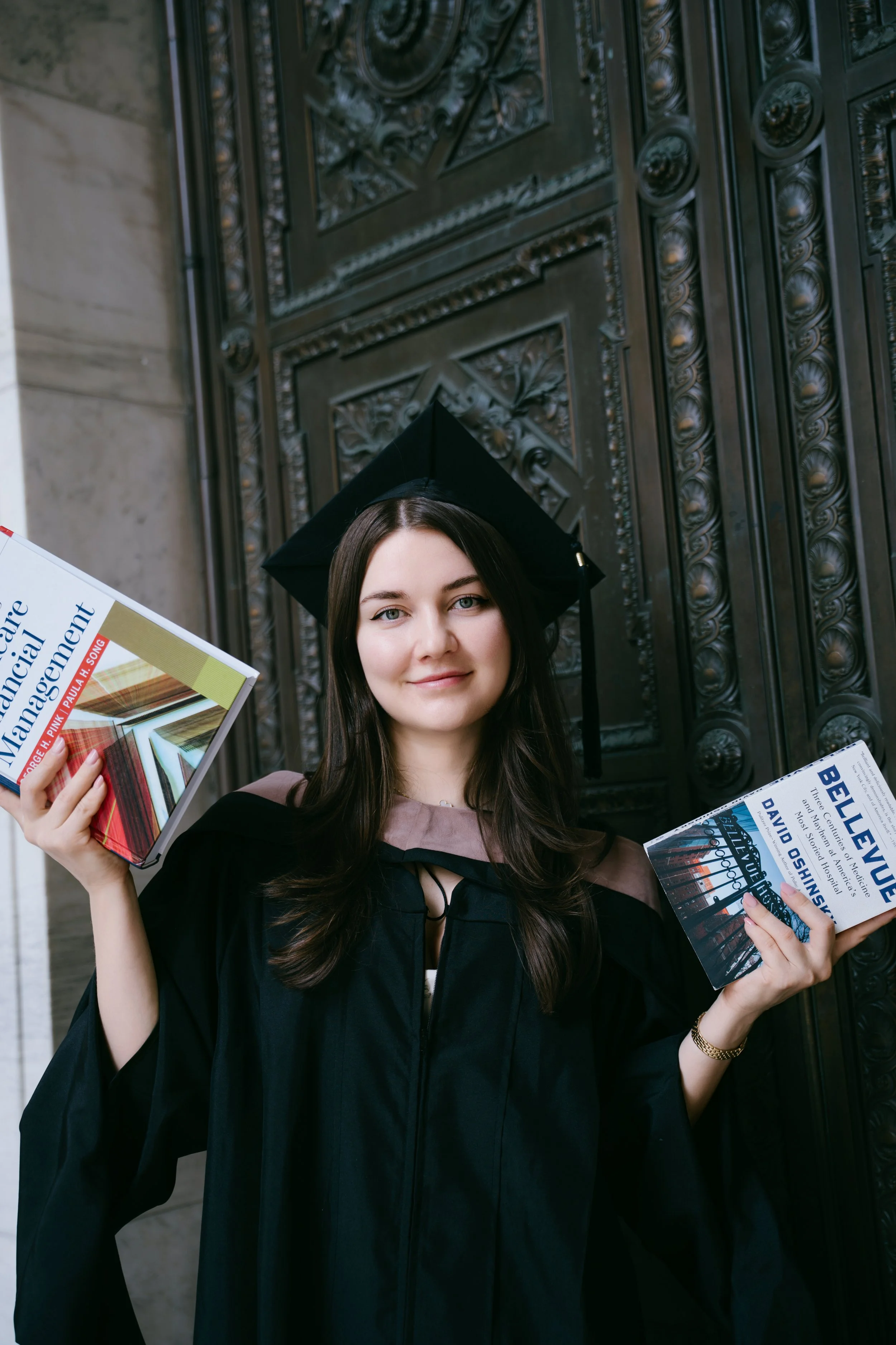 A young woman in graduation attire and cap, smiling and holding two books, one in each hand, standing in front of an ornate metal door.