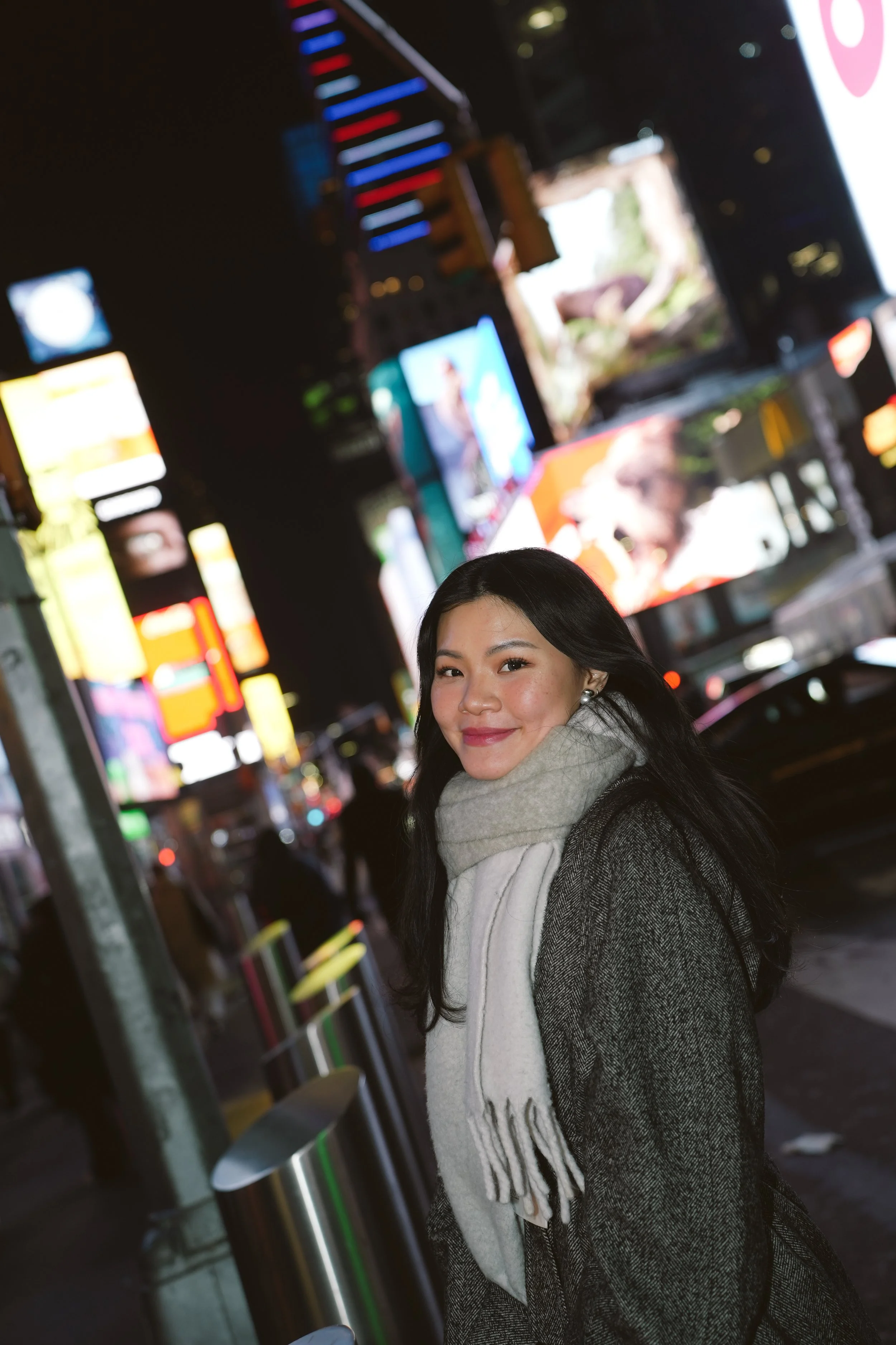 A woman with dark hair smiling in a city at night, standing near a parking ticket machine with colorful lights and digital screens in the background.