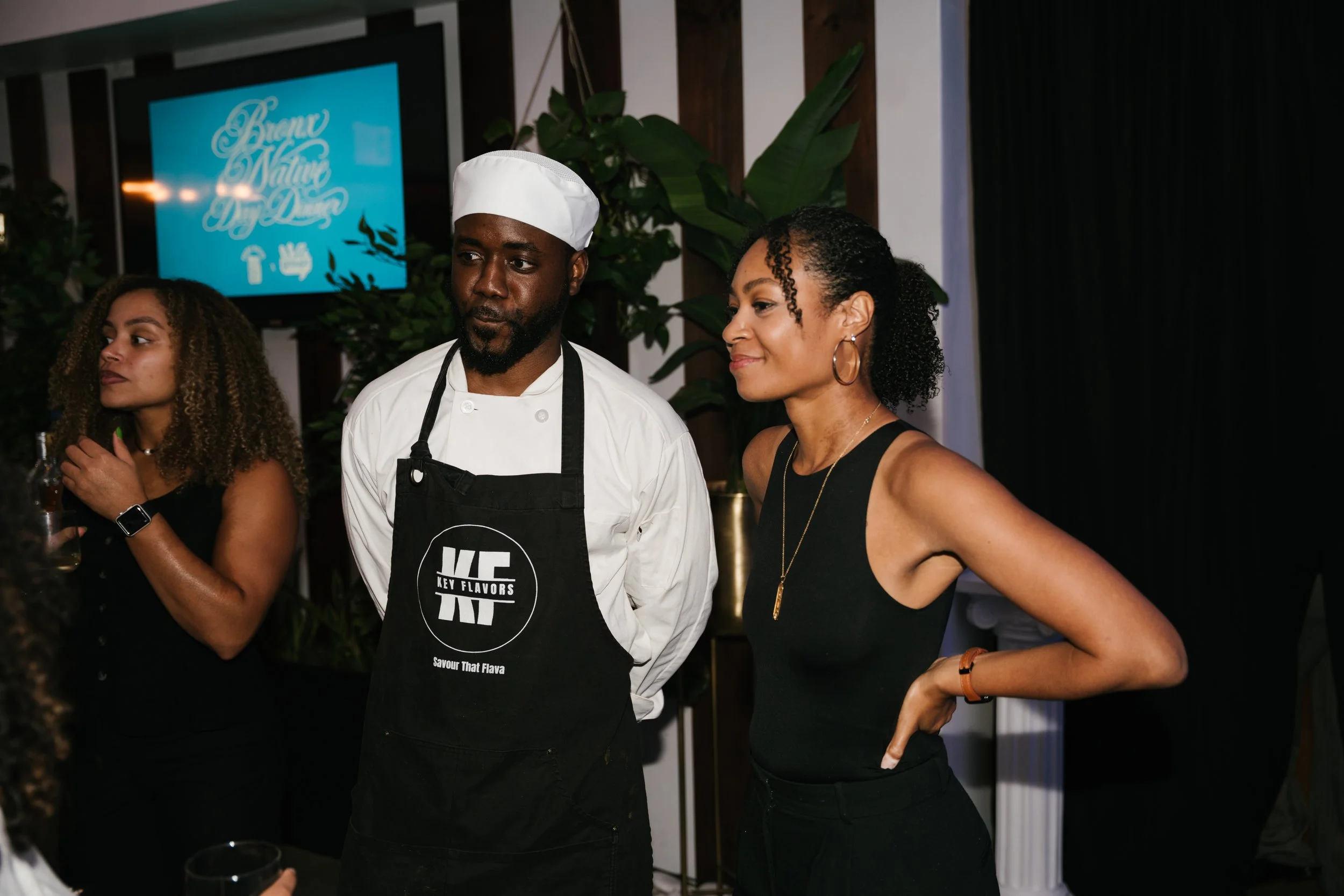 Three people standing together at an indoor event, with two women and a man dressed in a chef's uniform and apron, and a woman wearing hoop earrings and a black sleeveless top, engaged in conversation.