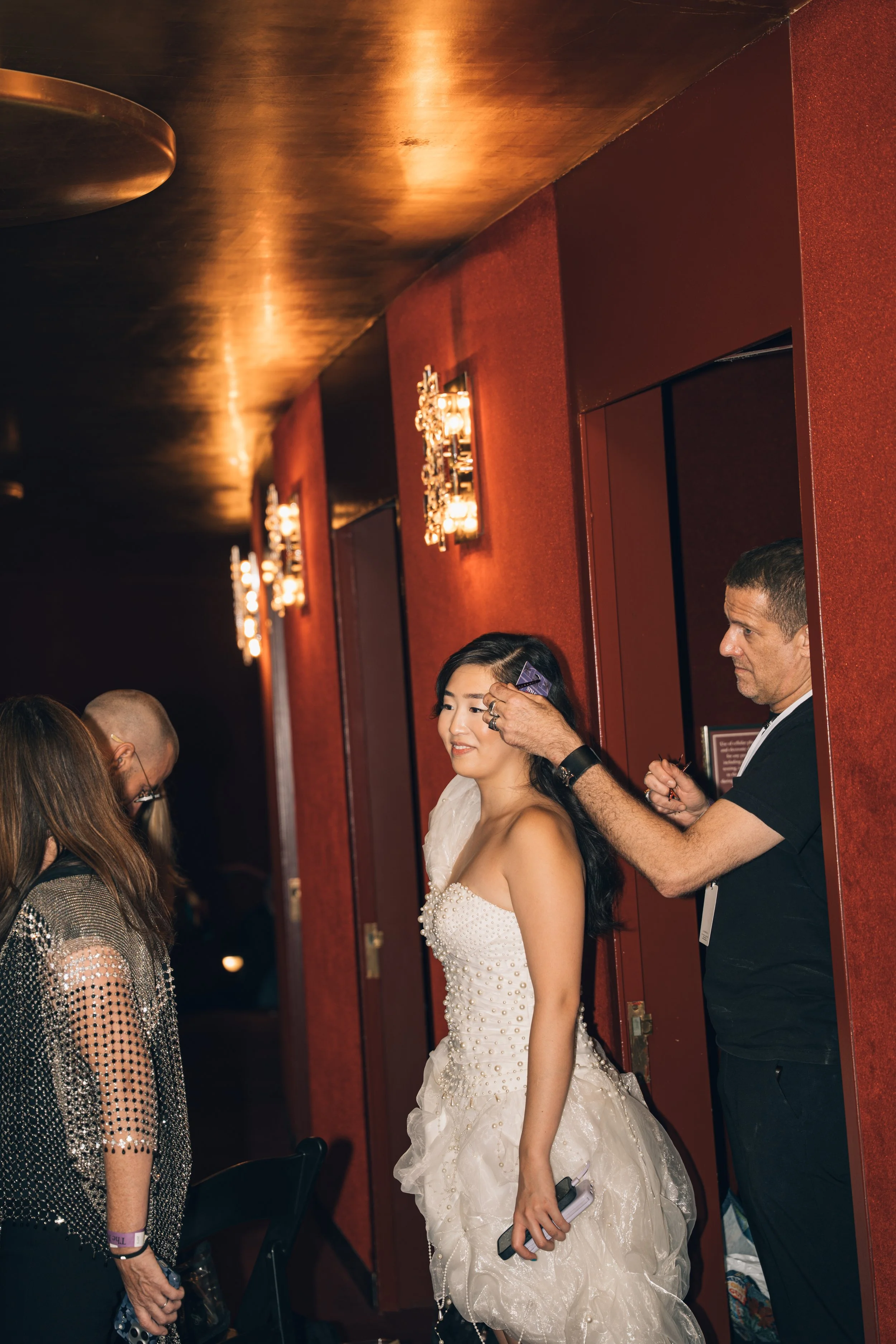 A woman in a white wedding dress receives a hairstyling or makeup touch-up from a man dressed in black, in a red-walled room with ornate light fixtures. Several other people are present, including a woman with long hair on the left and a man with gla