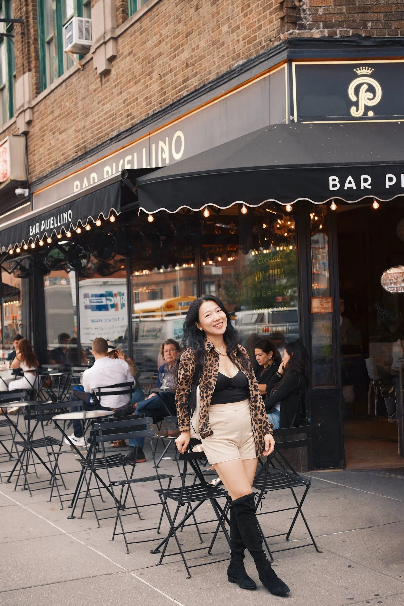 A woman standing in front of an outdoor restaurant, smiling. She is wearing a leopard print jacket, black top, beige shorts, and black boots, with a crowd of people dining and a restaurant with a black awning in the background.
