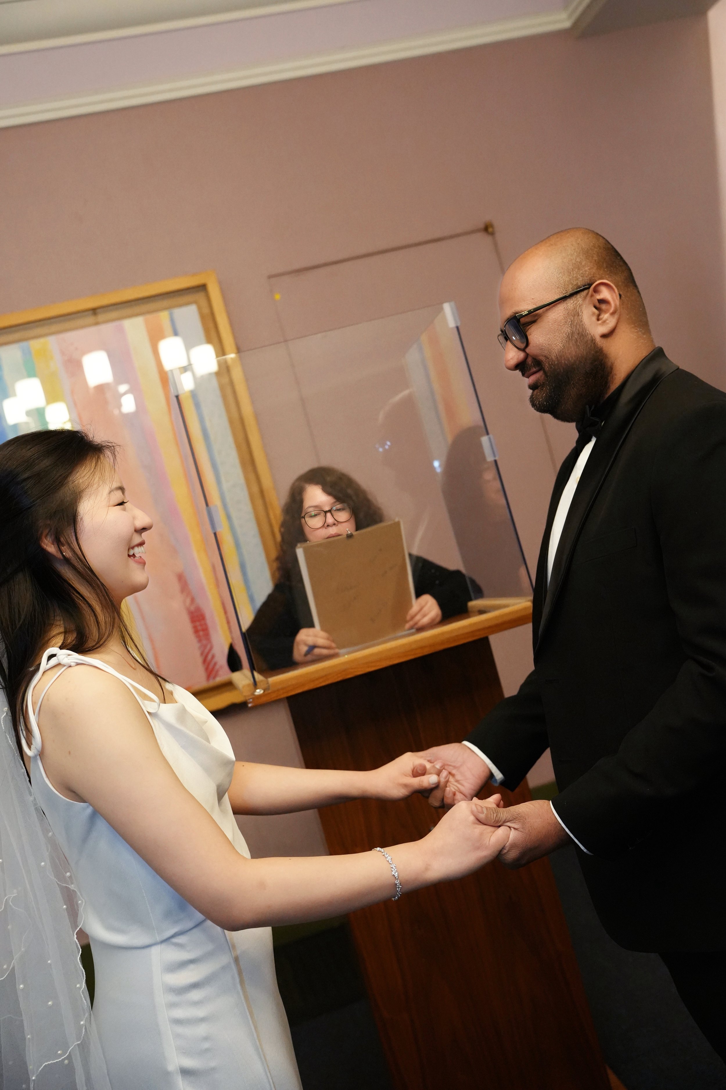A woman in a wedding dress and a man in a tuxedo shaking hands during a wedding ceremony while a woman at a podium looks on.
