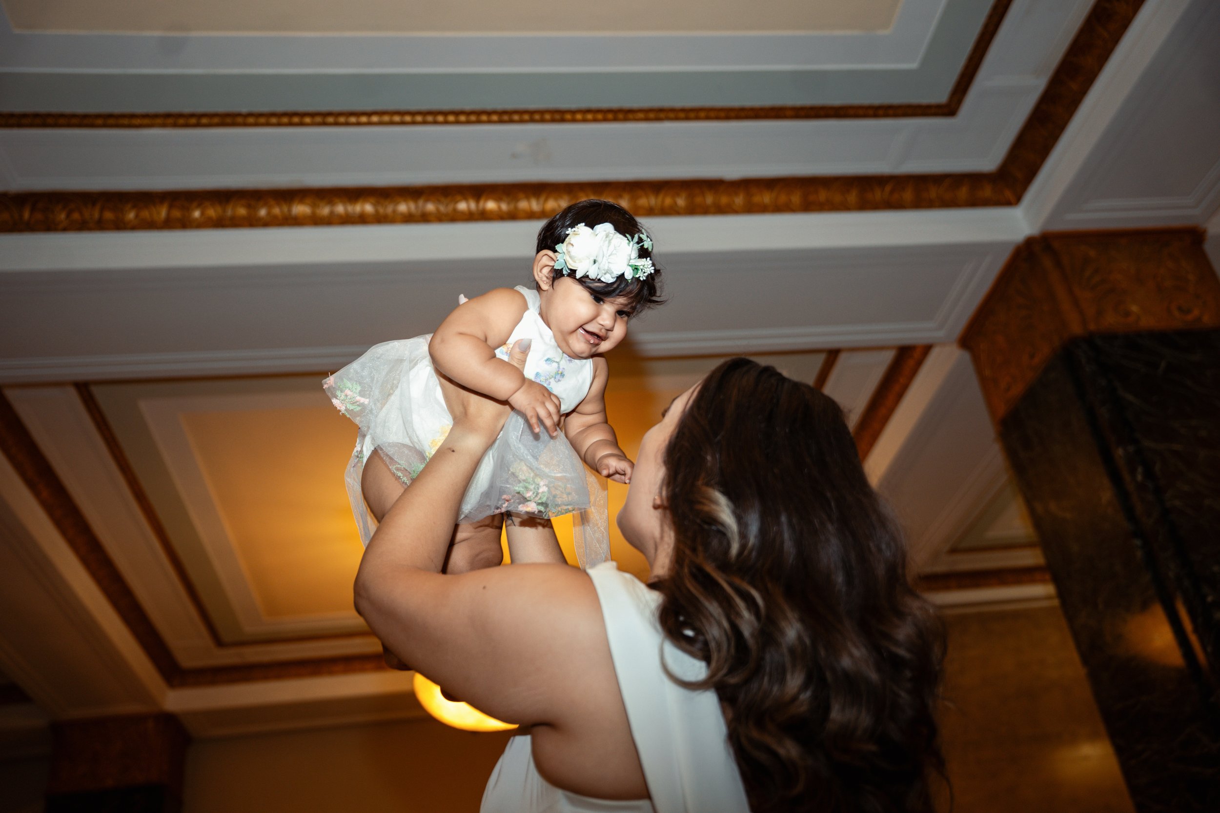 A woman lifting a young girl dressed in a white dress with floral details and a flower headband, in a room with ornate ceiling moldings and warm lighting.