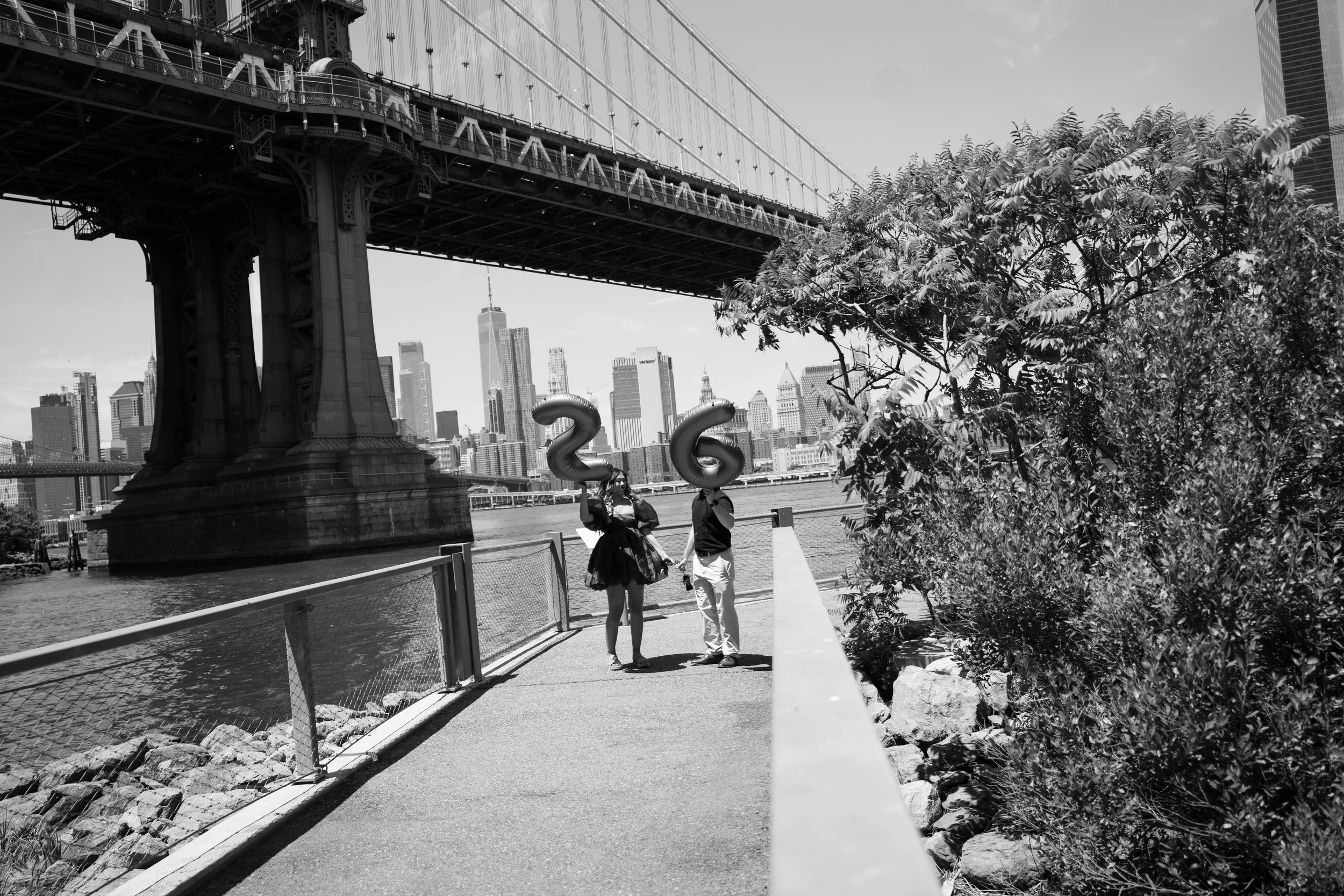 A black-and-white photograph of the Brooklyn Bridge with the New York City skyline in the background. Two people on a riverside walkway hold balloon numbers '2' and '6'. The scene includes trees and fencing along the path.