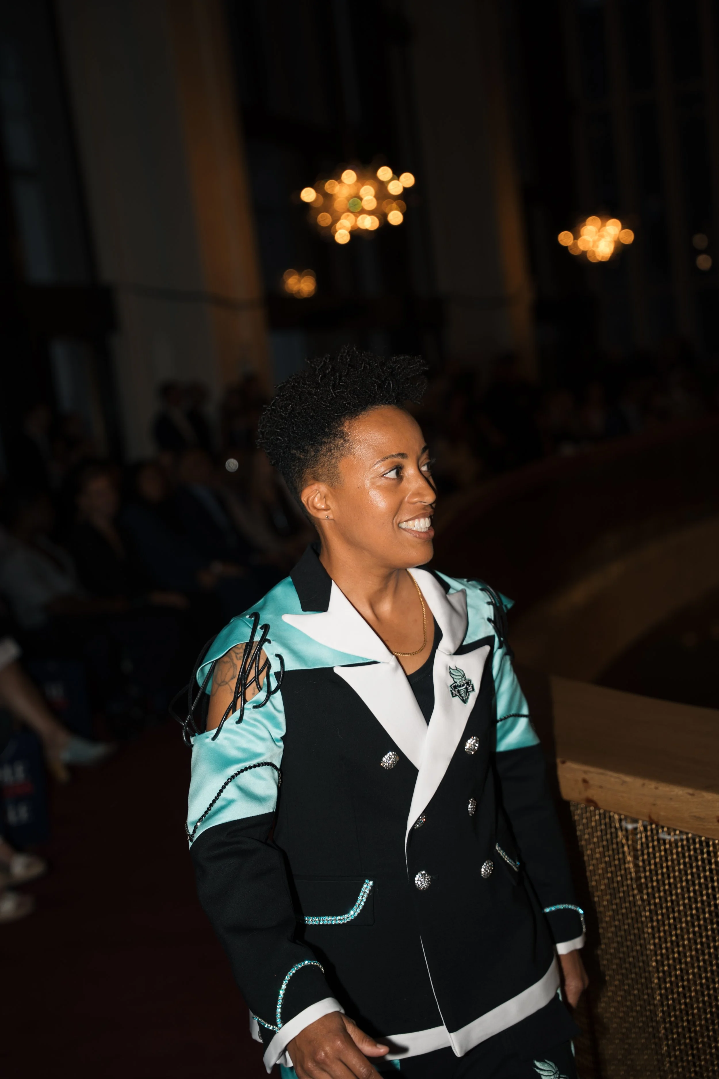 A smiling woman with short curly hair wearing a black, white, and turquoise blazer with silver embellishments, standing in a dark room with chandeliers and other people in the background.