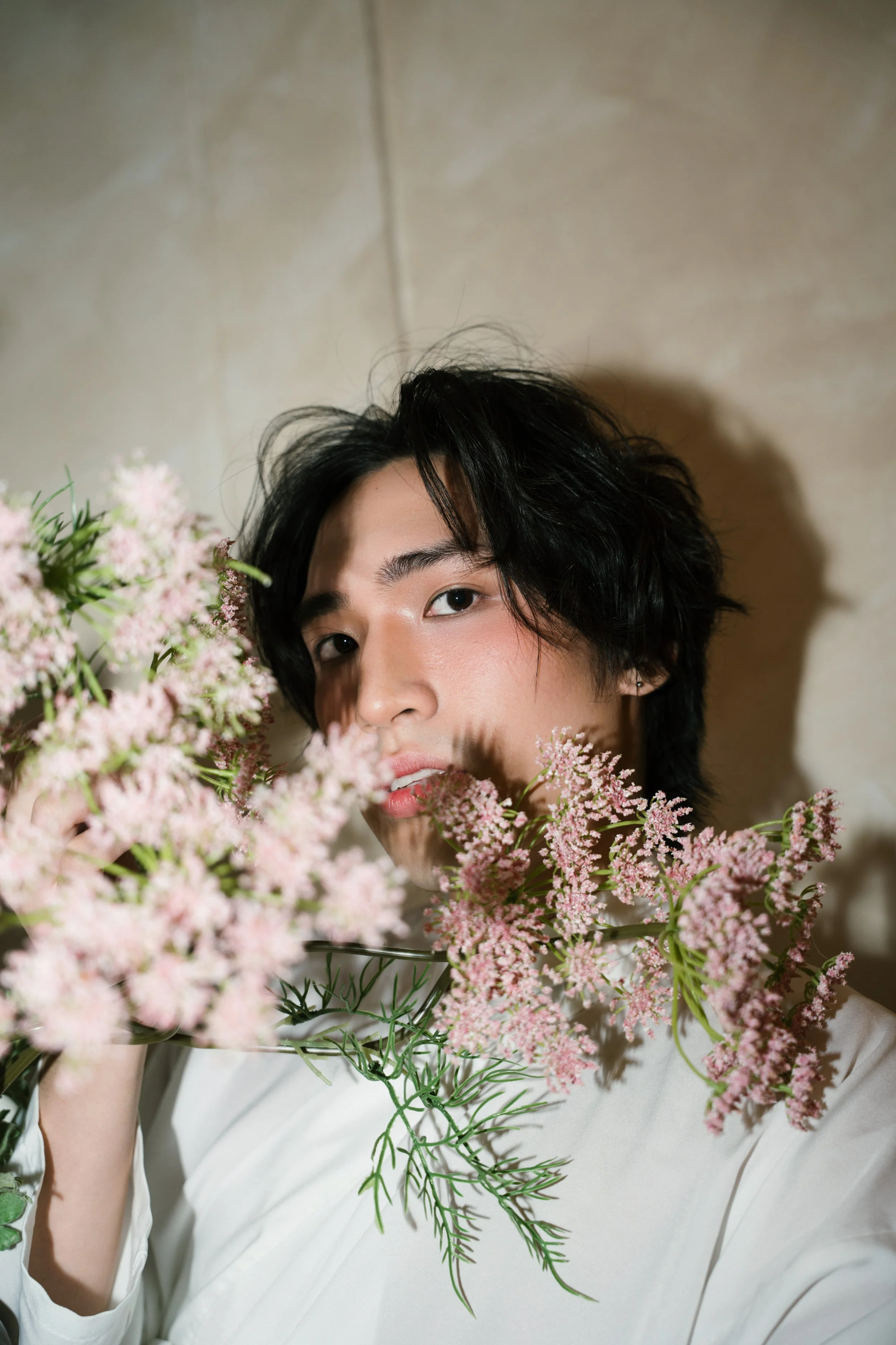 A person with black, wavy hair and light skin holding pink flowers in front of their face, wearing a white shirt.
