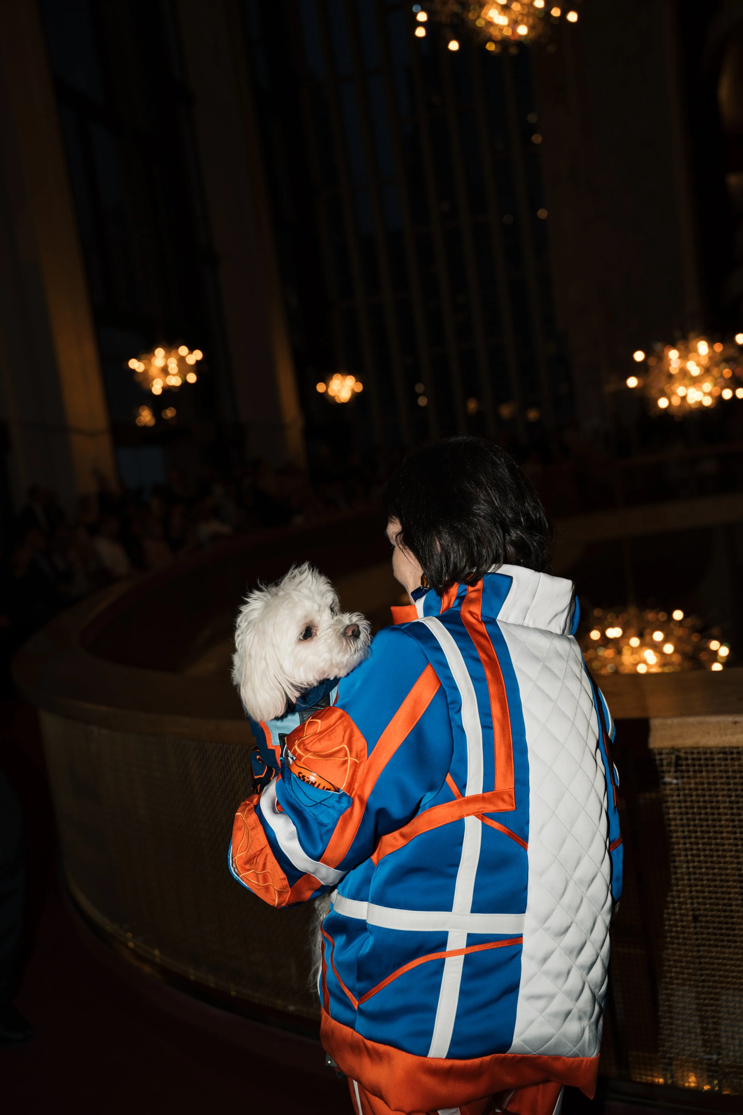 A woman with dark hair wearing a colorful jacket holding a small white dog in her arms. The background features a circular glass window and blurred warm lights, suggesting an indoor setting with a cozy atmosphere.