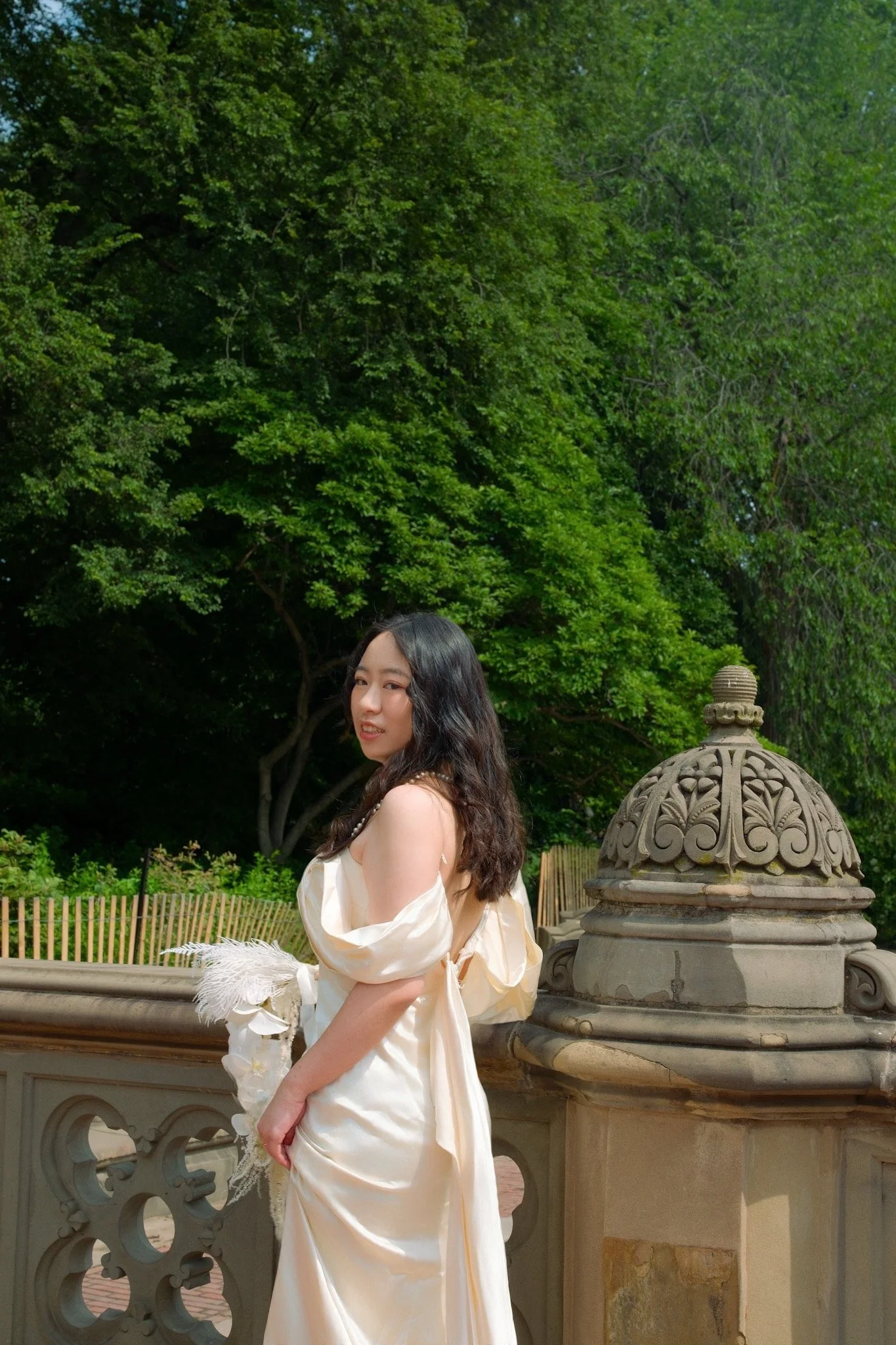 Young woman with long dark hair wearing a cream-colored dress, standing outdoors next to a decorative stone fountain with lush green trees in the background.