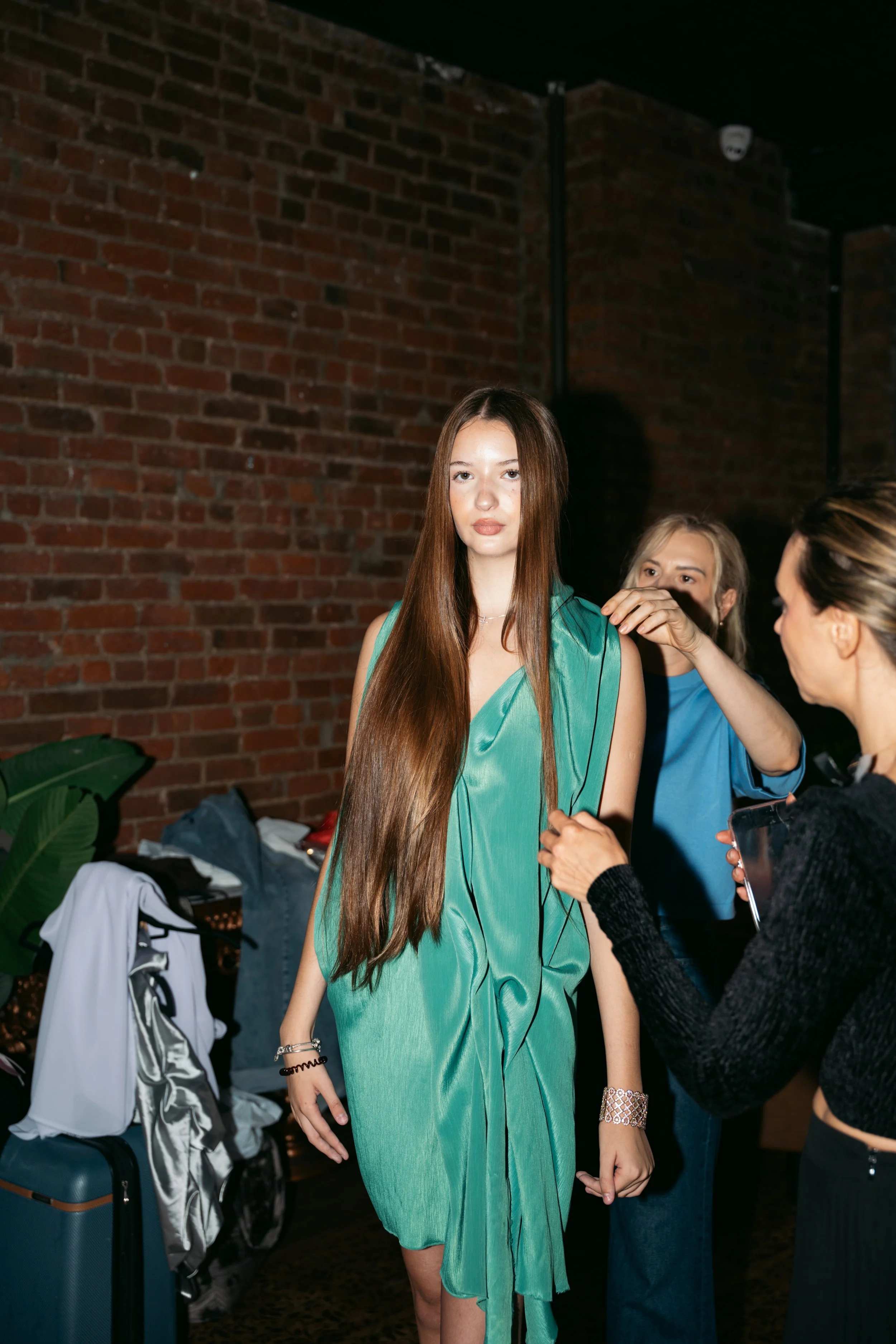 A young woman with long, straight brown hair and light skin is getting ready, with the help of two women behind her, in front of a brick wall. She is wearing a green dress and has jewelry on her wrists. The setting appears to be backstage or in a dre
