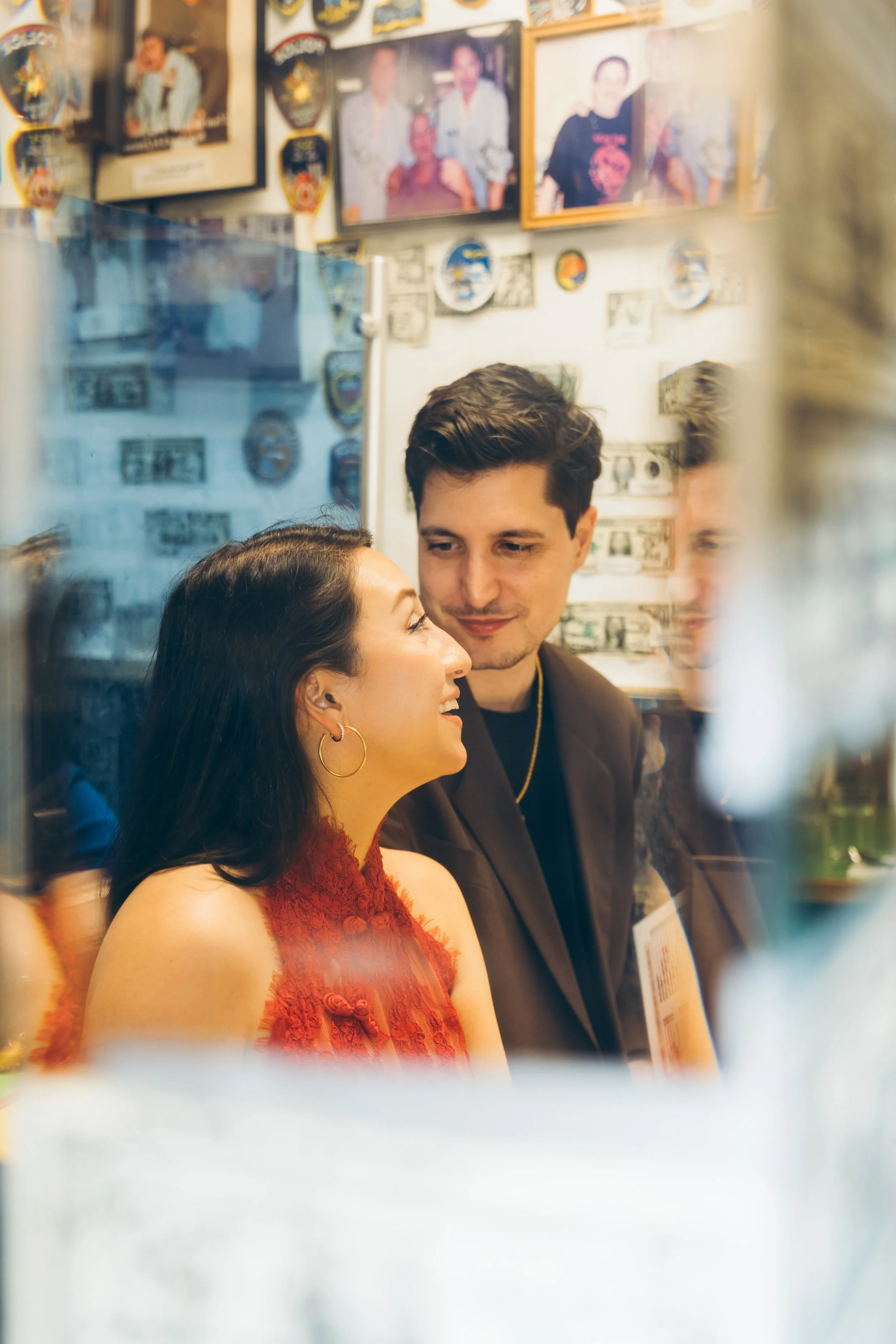 A young woman with dark hair wearing gold hoop earrings and a red sleeveless dress smiling and looking at a young man with dark hair dressed in a dark suit. They are inside a room decorated with framed photos, patches, and dollar bills on the wall, s