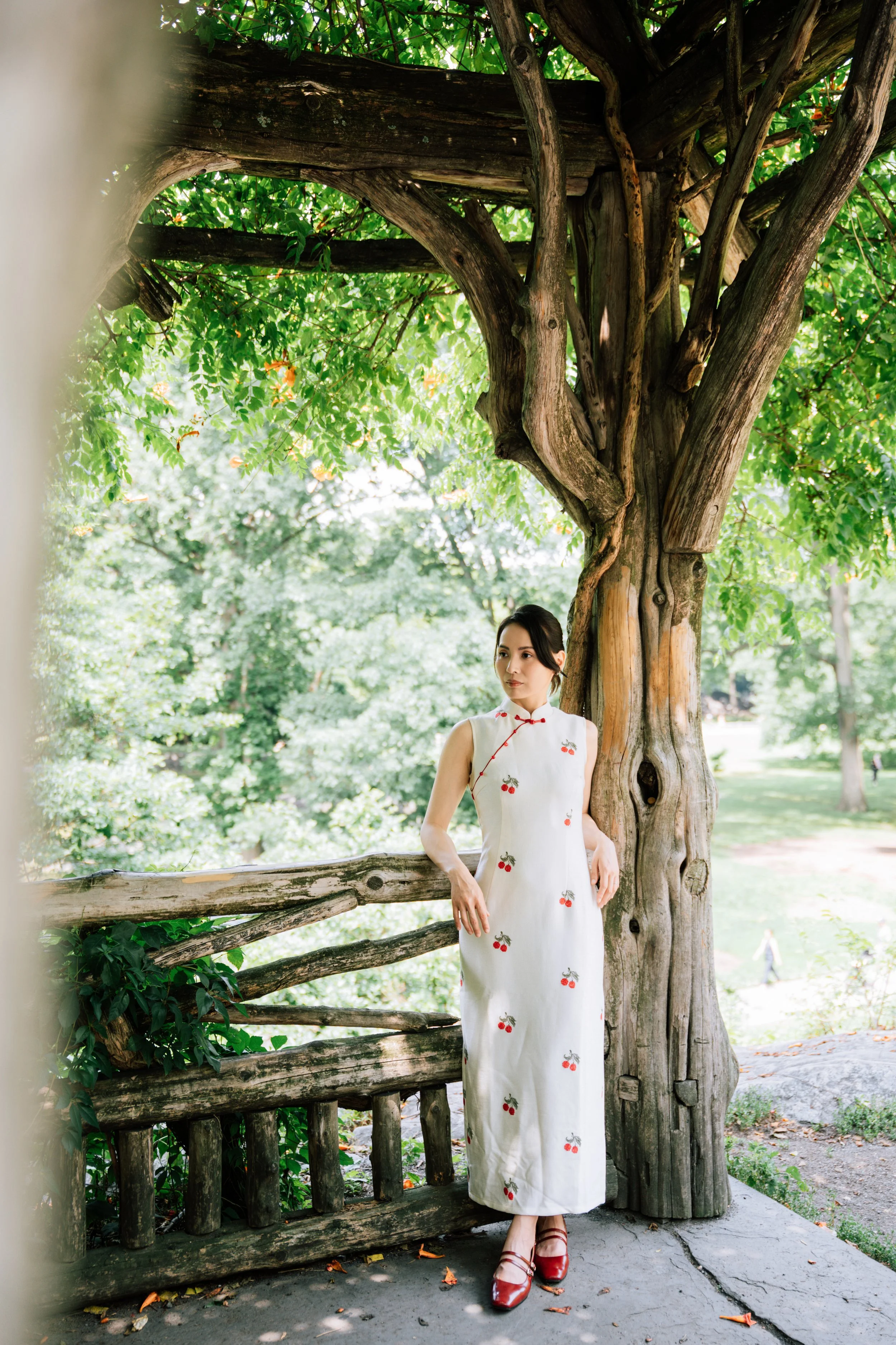 A woman in a white dress with cherry print standing under a large tree with twisted branches and a wooden fence, in a park with green foliage in the background.