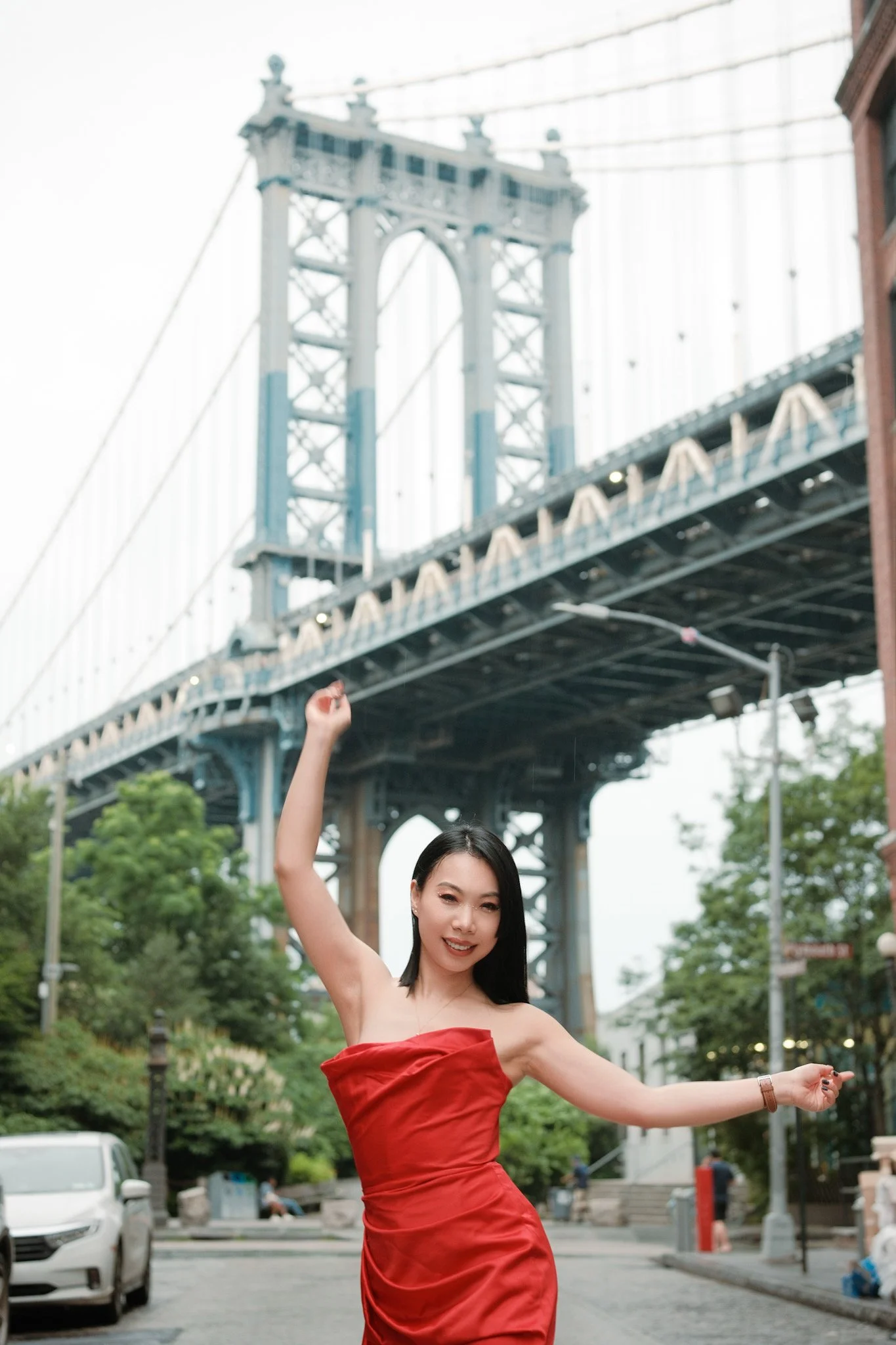 A woman in a red dress dancing outdoors near the San Francisco Bay Bridge with arms raised, smiling, surrounded by trees and parked cars.