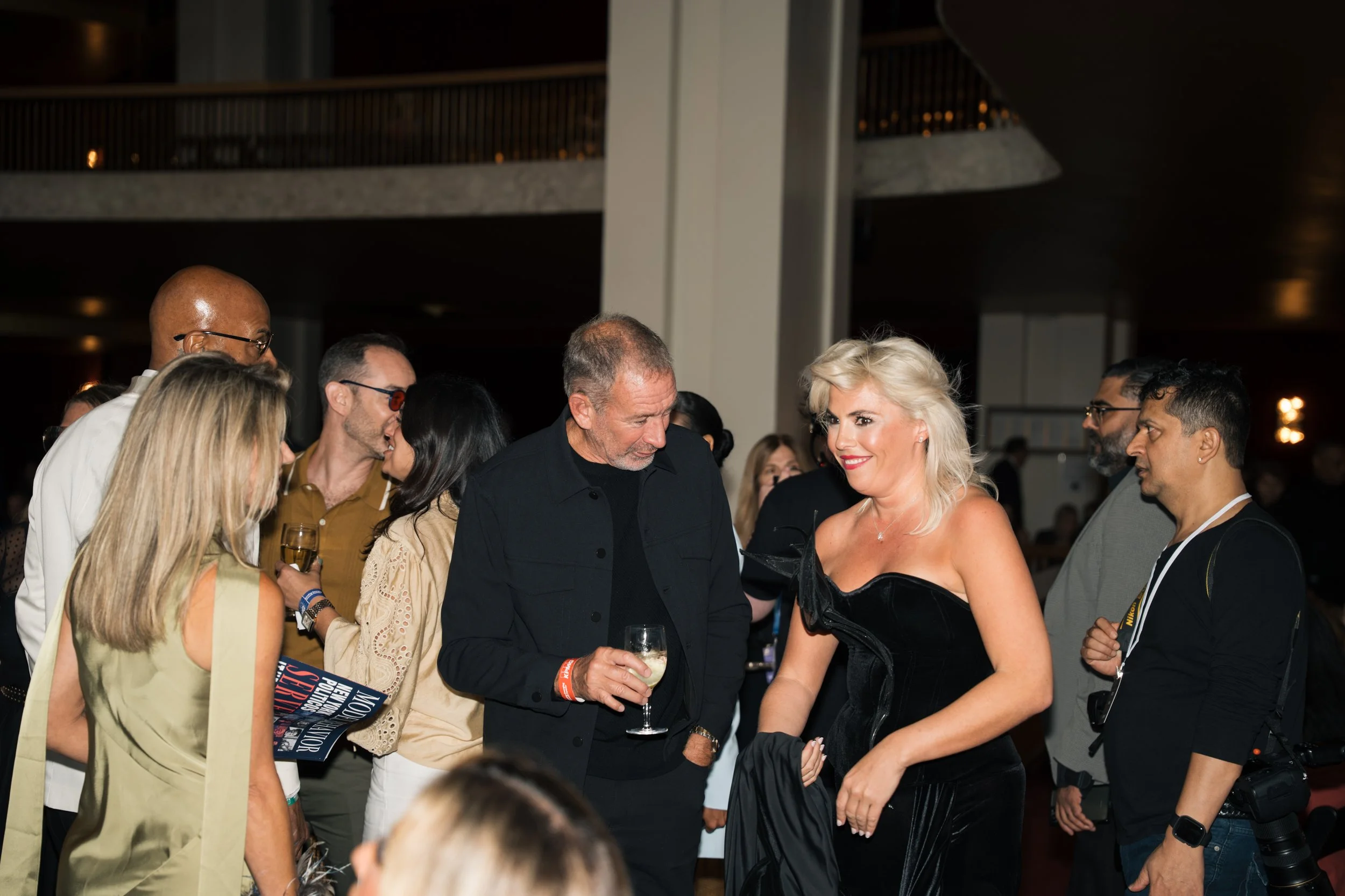 Group of well-dressed people socializing at an indoor event, including a woman in a black dress receiving a drink from a man, with others mingling in the background.