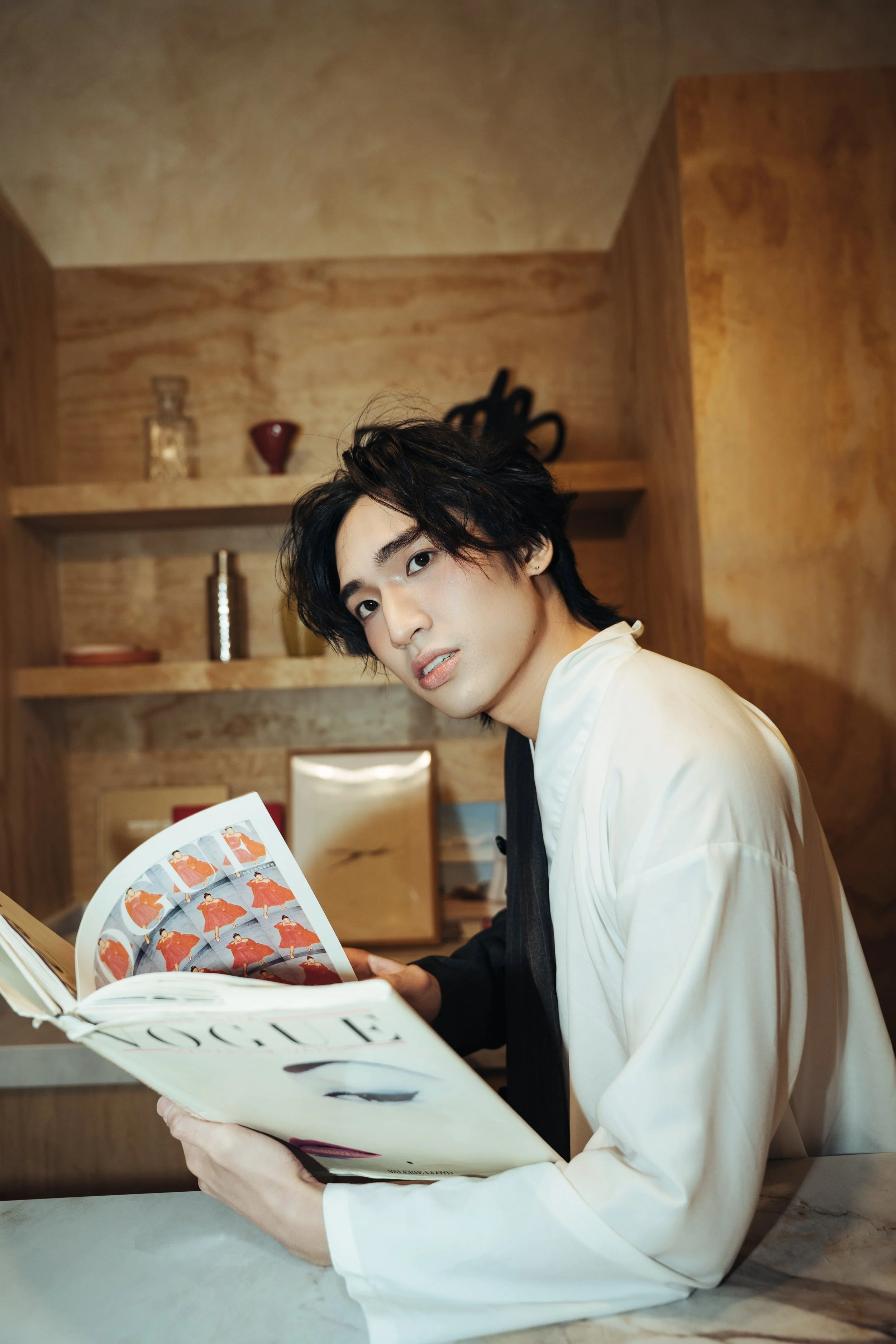 A young man with black, wavy hair and fair skin is sitting at a table, looking at a magazine with fashion images. He is wearing a white shirt with rolled-up sleeves and appears to be in a cozy, wood-paneled room with shelves containing decorative vas