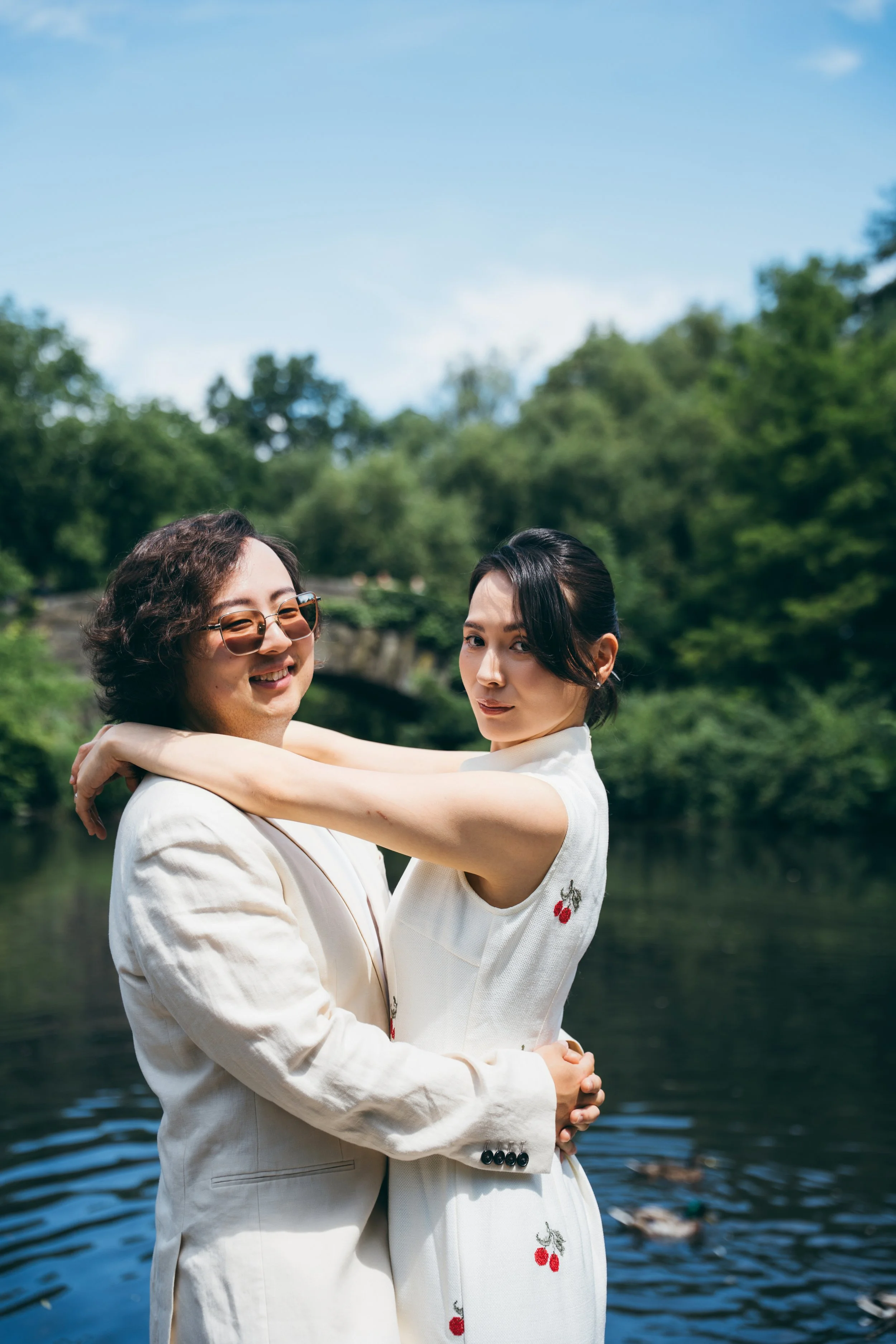A man and woman standing close together by a river with lush greenery in the background, embracing and smiling at the camera.