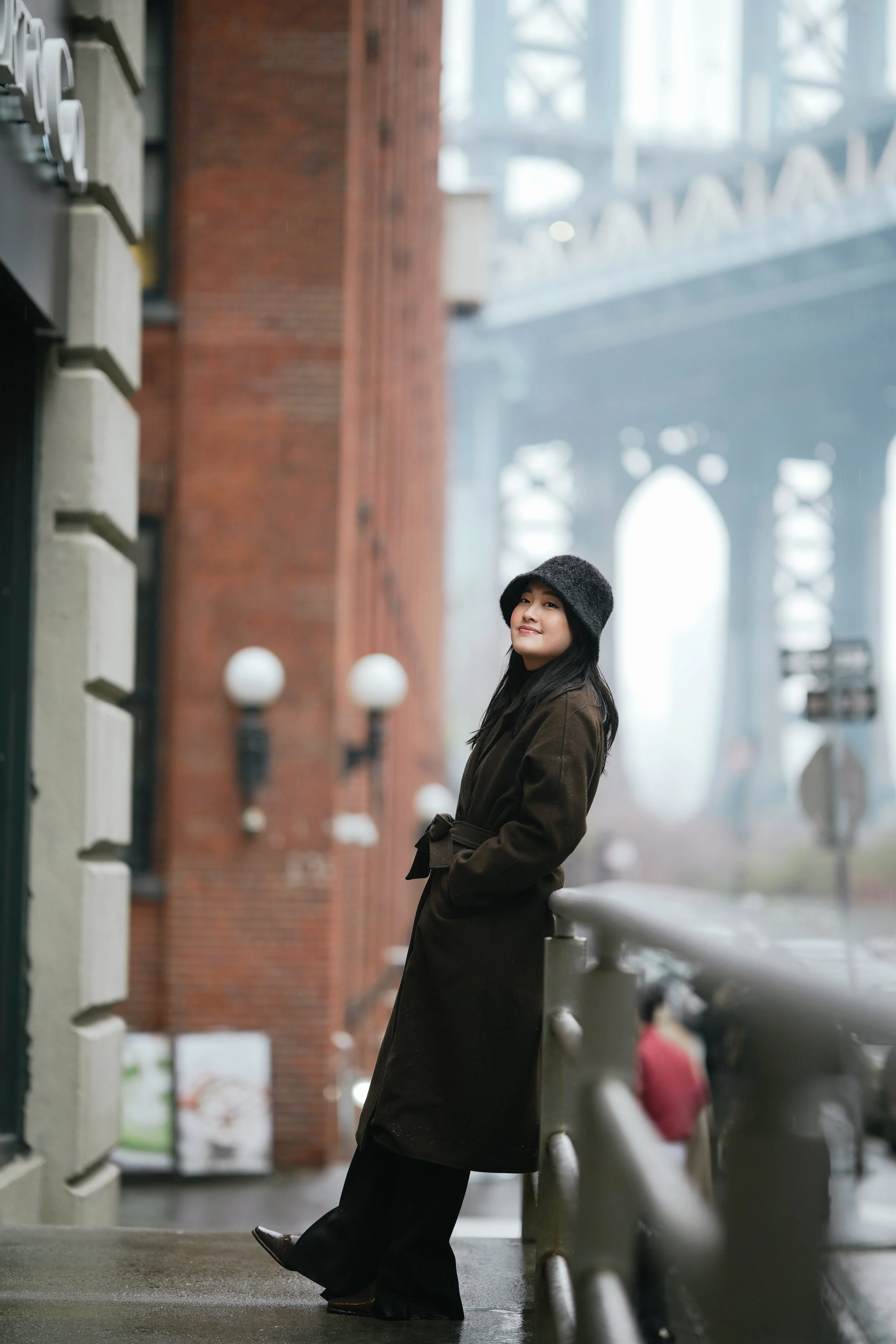 A woman with long dark hair wearing a black hat and brown coat, standing on a city street, leaning on a white railing, smiling slightly, with blurred background of buildings and other people.
