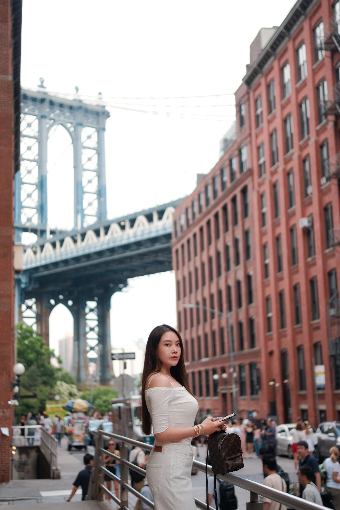 A young woman with long dark hair standing on a city street, holding a phone and a Louis Vuitton bag, with the Brooklyn Bridge and red brick buildings in the background.