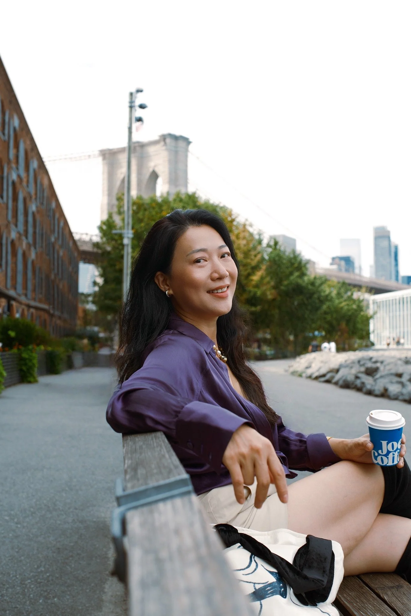 A woman with long dark hair and hoop earrings smiling while sitting on a park bench with a coffee cup in her hand, in an urban setting with trees, buildings, and a bridge in the background.