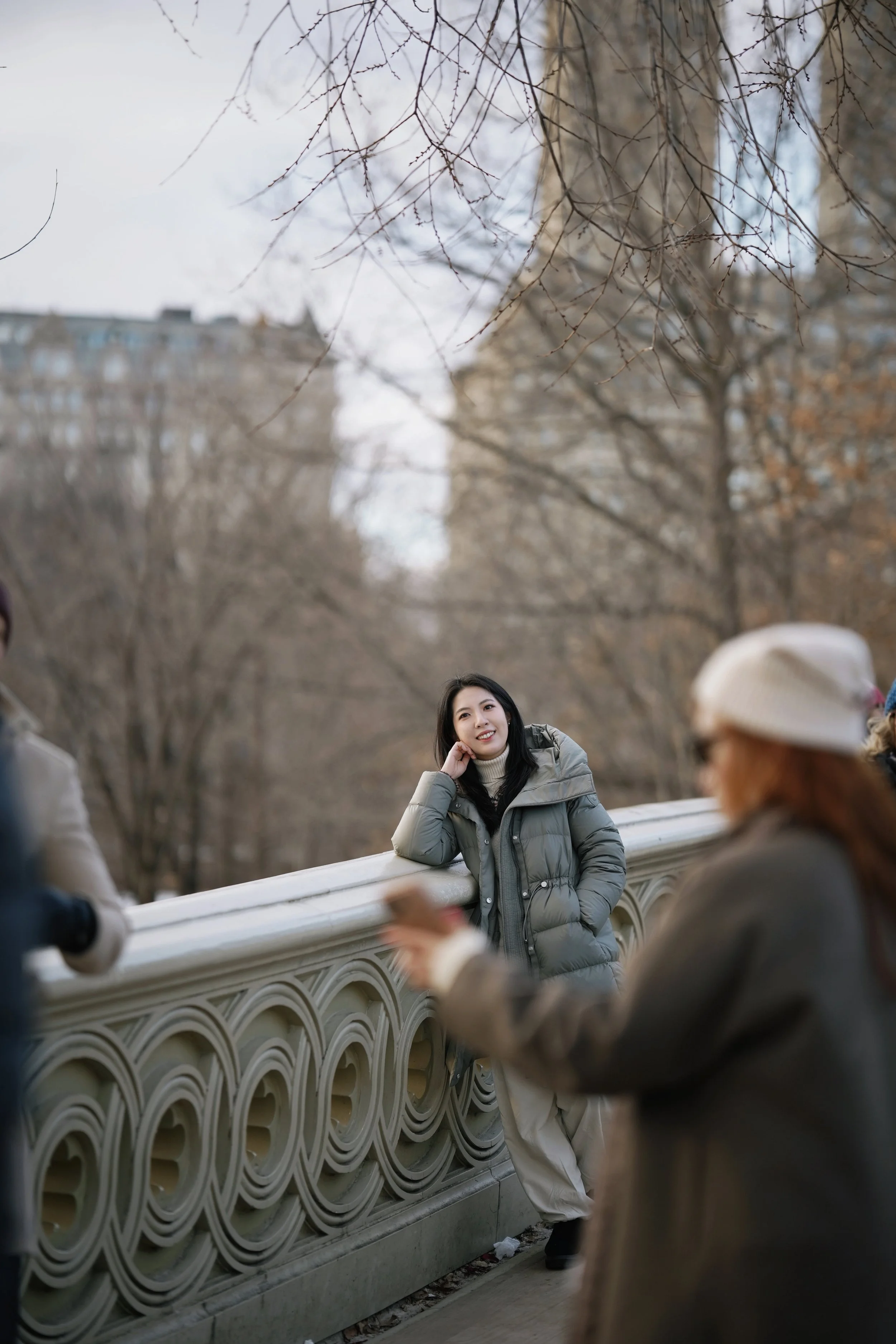 A woman in a gray puffer jacket leaning on a decorative bridge railing, looking at two older women who are talking nearby, with leafless trees and buildings in the background on a cloudy day.
