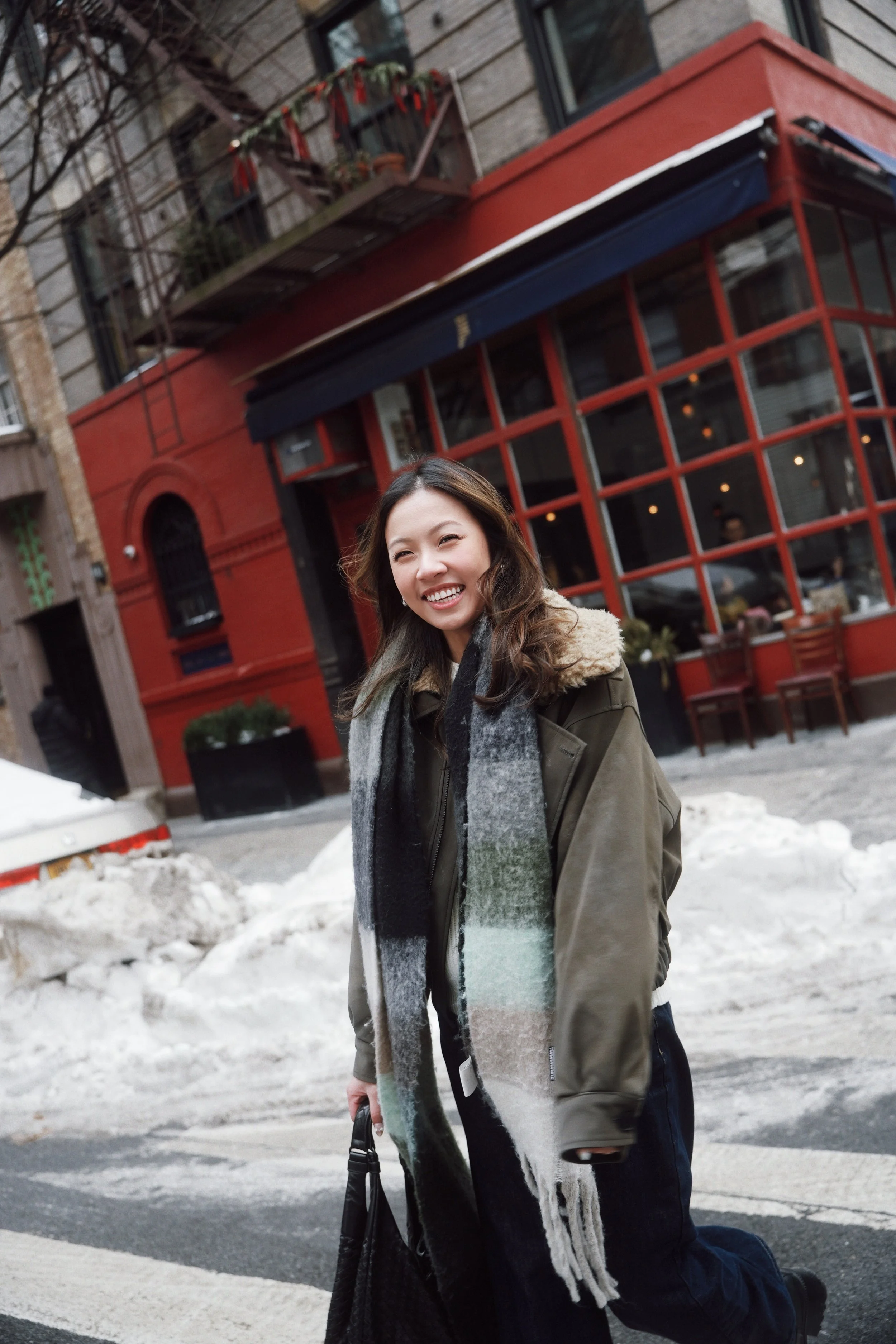 A smiling woman with long brown hair, wearing a green jacket and a multicolored scarf, kneeling on a street with snow piles, in front of a red building with large windows, during winter.