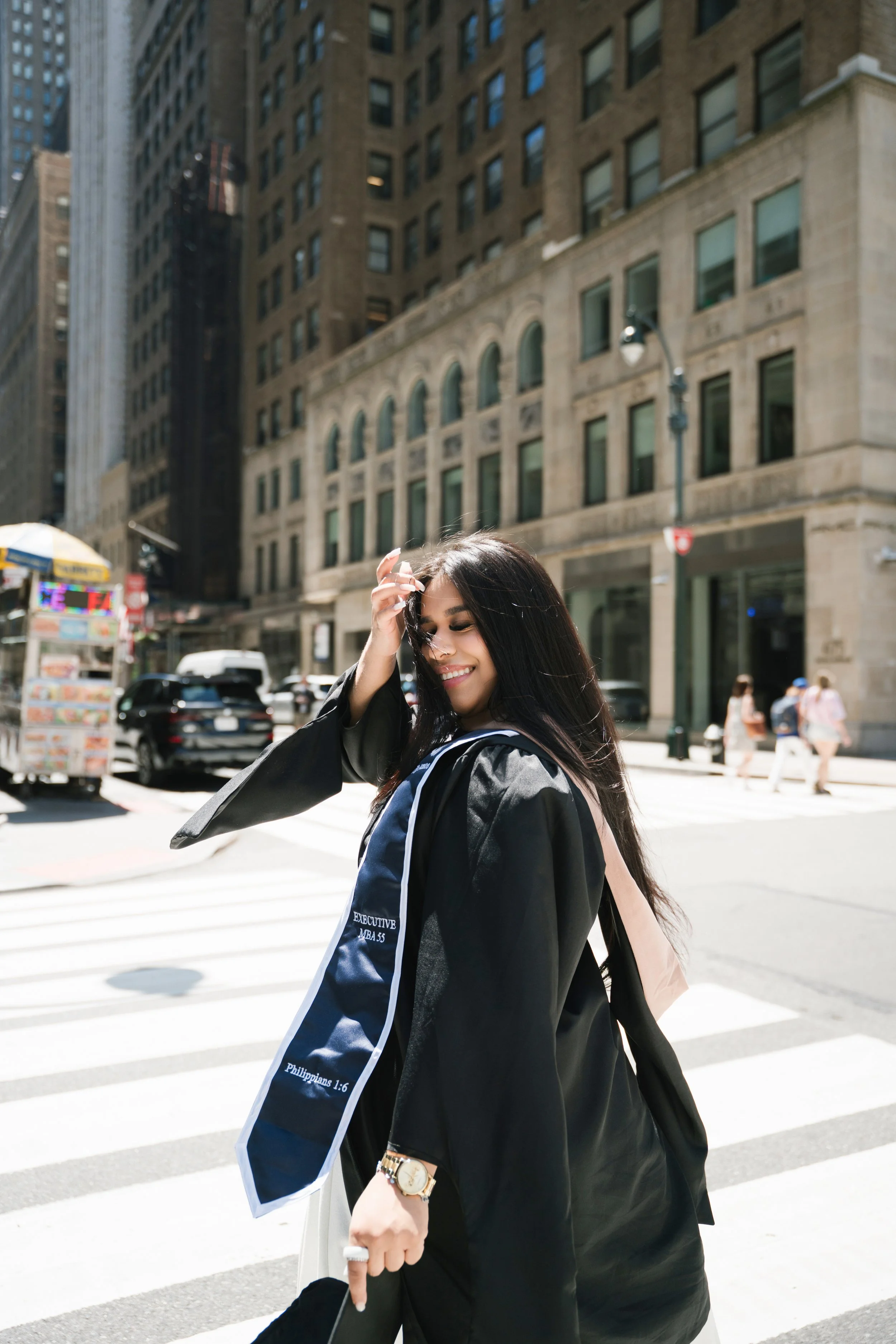 A smiling young woman with long dark hair, wearing a black graduation hoodie with the words 'EXECUTIVE MBA 55' and a Bible verse from Philippians 1:6, is crossing a city street. She is touching her hair with one hand and carrying a bag and a cell pho