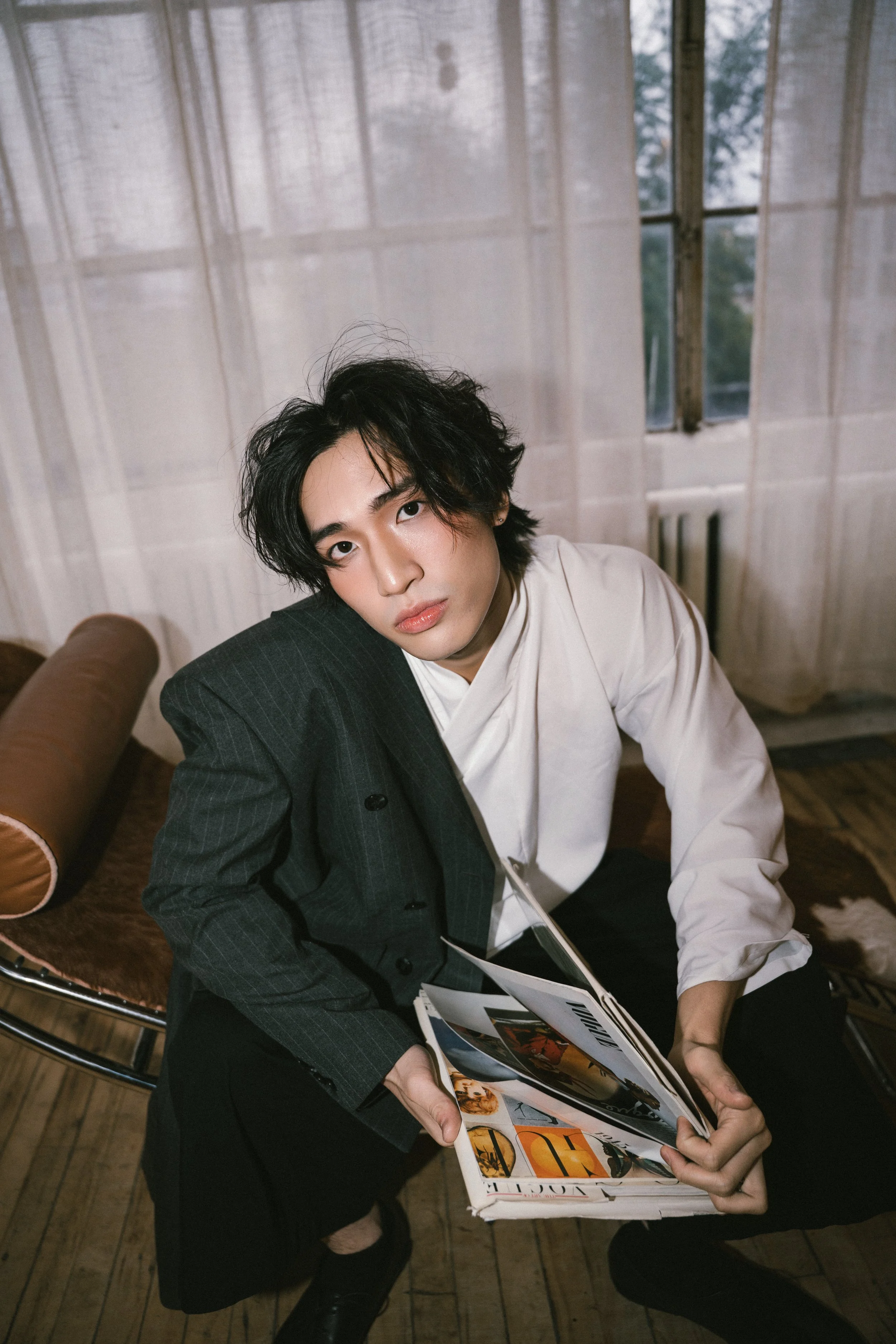 A young man with black wavy hair sitting in a room with large windows and sheer curtains, wearing a black pinstripe blazer over a white shirt, holding an open magazine and looking at the camera.