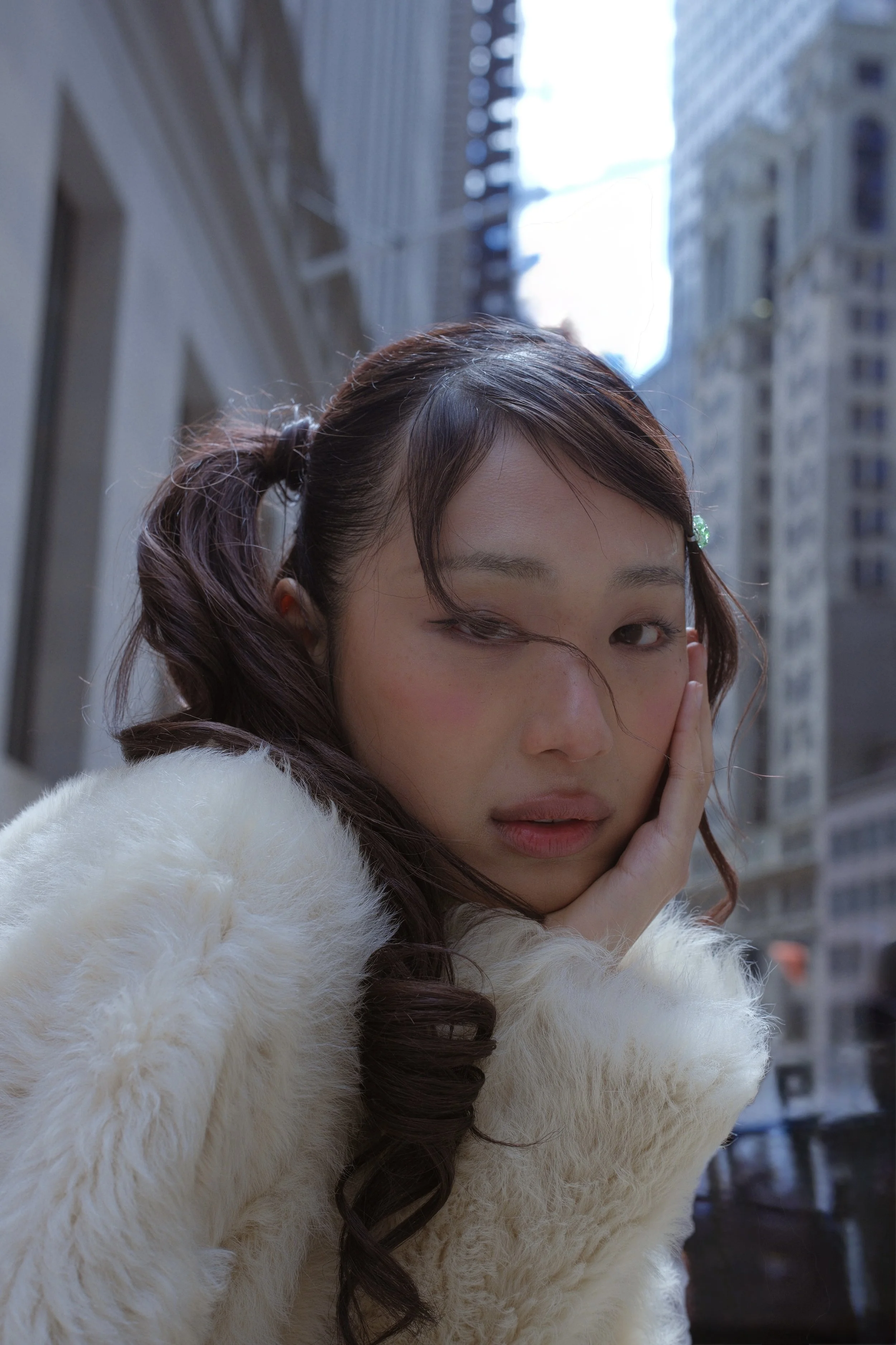 A young woman with brown hair styled in a loose braid, wearing a white faux fur coat, is resting her face in her hand and looking at the camera with a city street and tall buildings in the background.