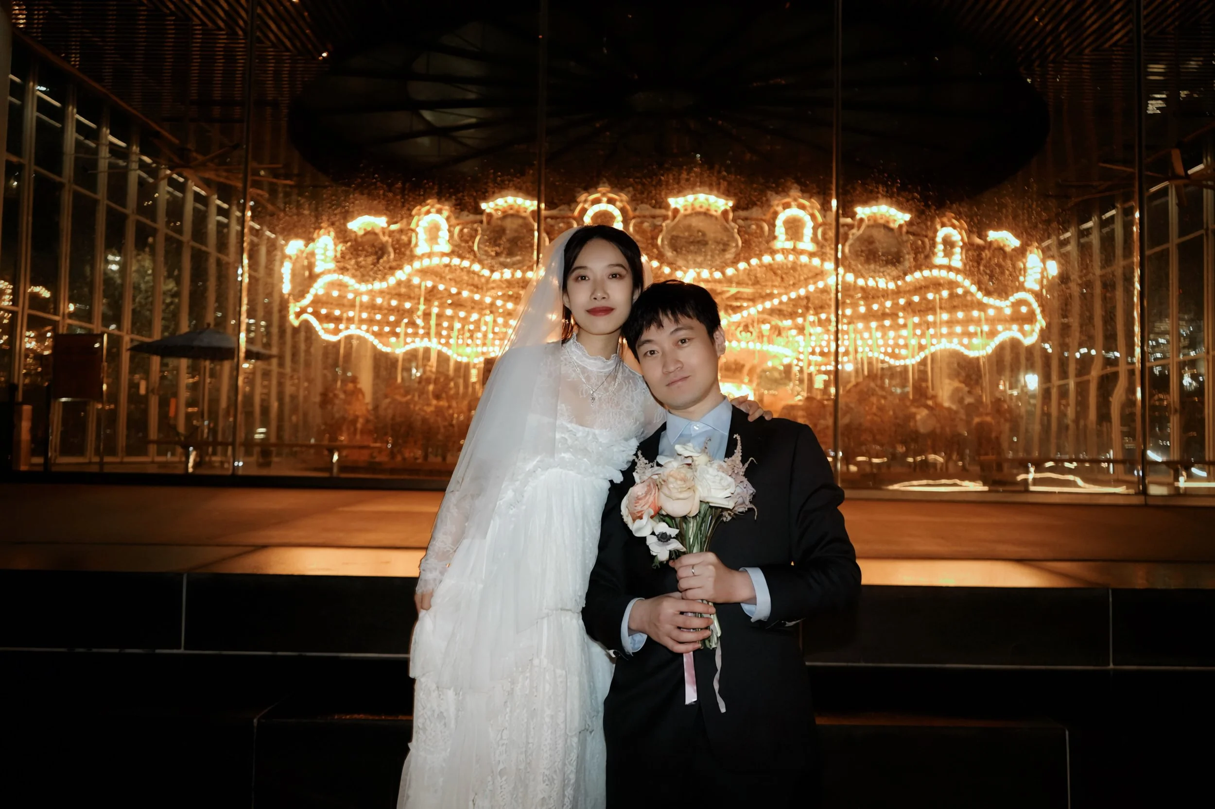 A bride and groom posing in front of a carousel with bright lights at night. The bride is wearing a white wedding dress and veil, while the groom is dressed in a black tuxedo holding a bouquet of roses.