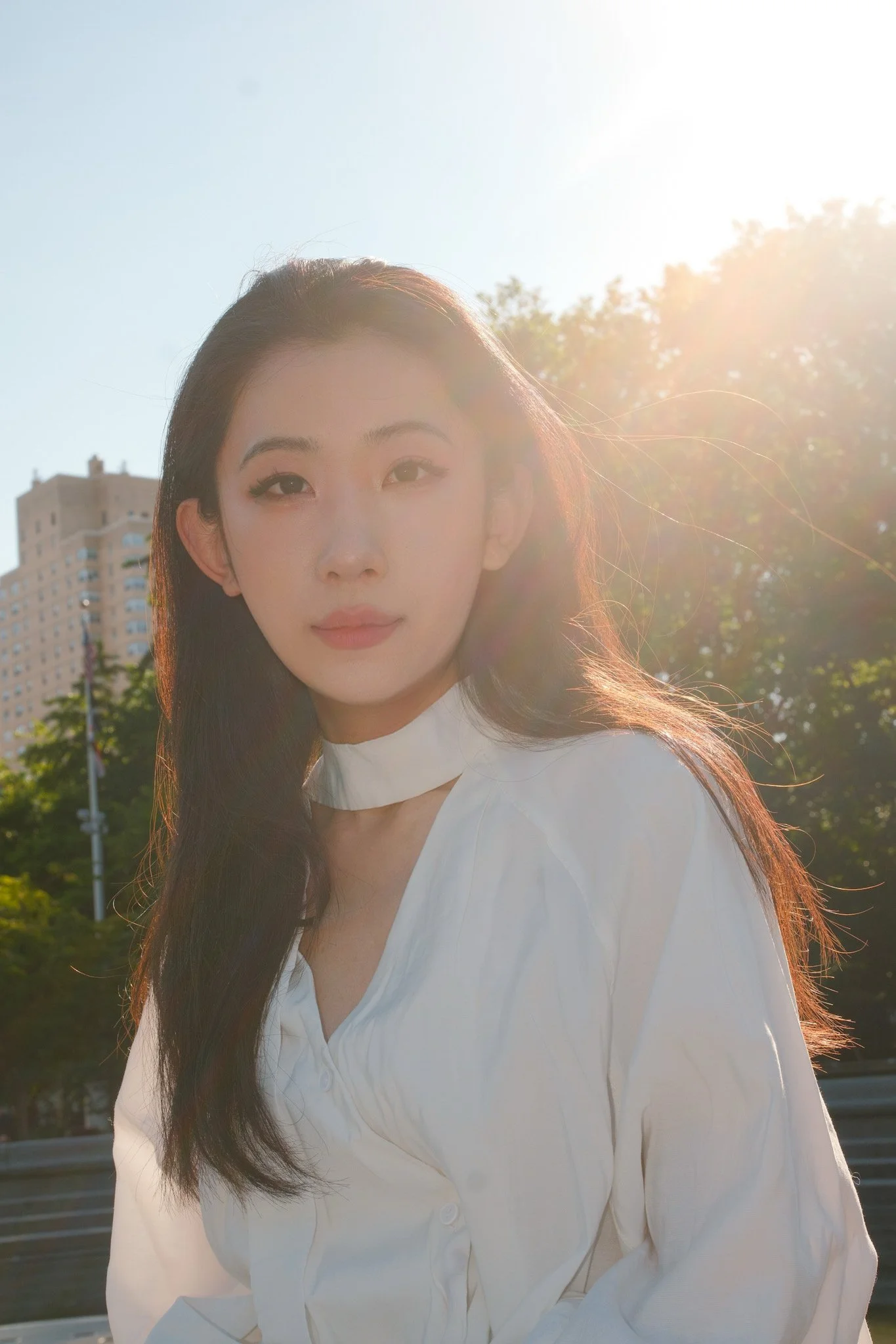 A young woman with long dark hair and a light complexion standing outdoors in front of trees and a building, with sunlight shining from behind her.