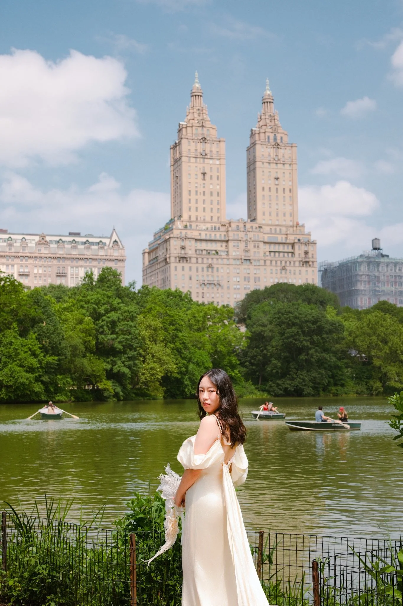 A woman in a white dress standing by a lake with boats, lush green trees, and a city skyline with tall buildings and blue sky in the background.