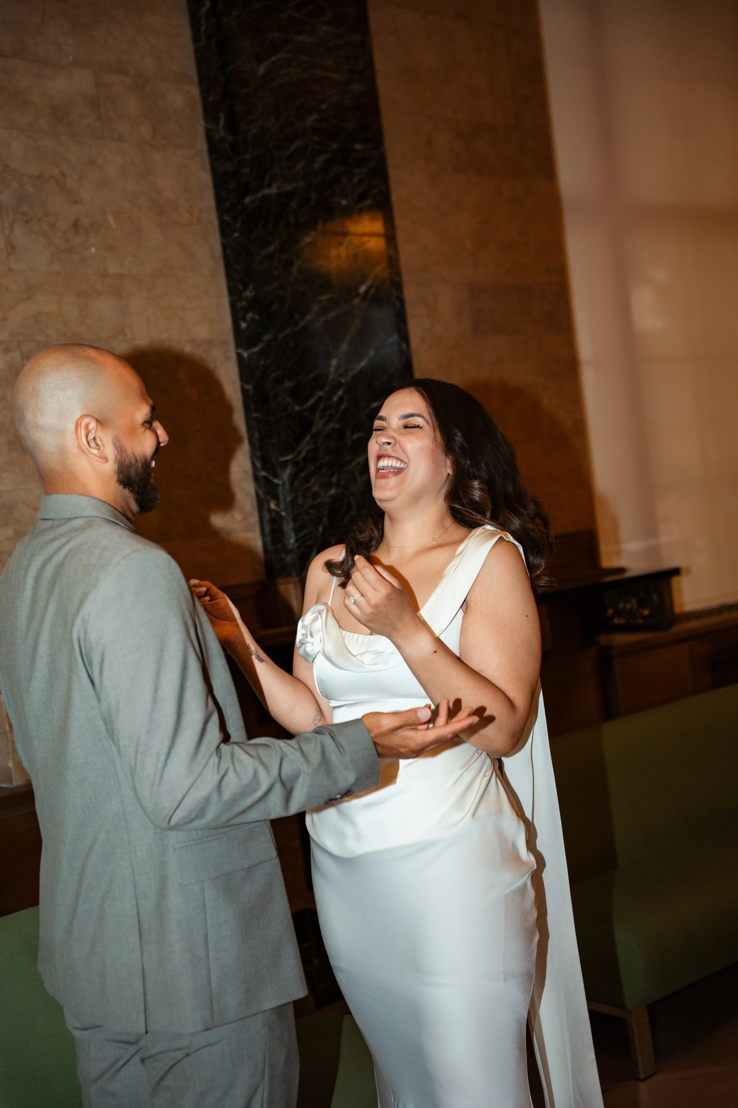 A woman in a white dress and a man in a gray suit sharing a joyful moment, holding hands and smiling at each other inside a building with marble walls.