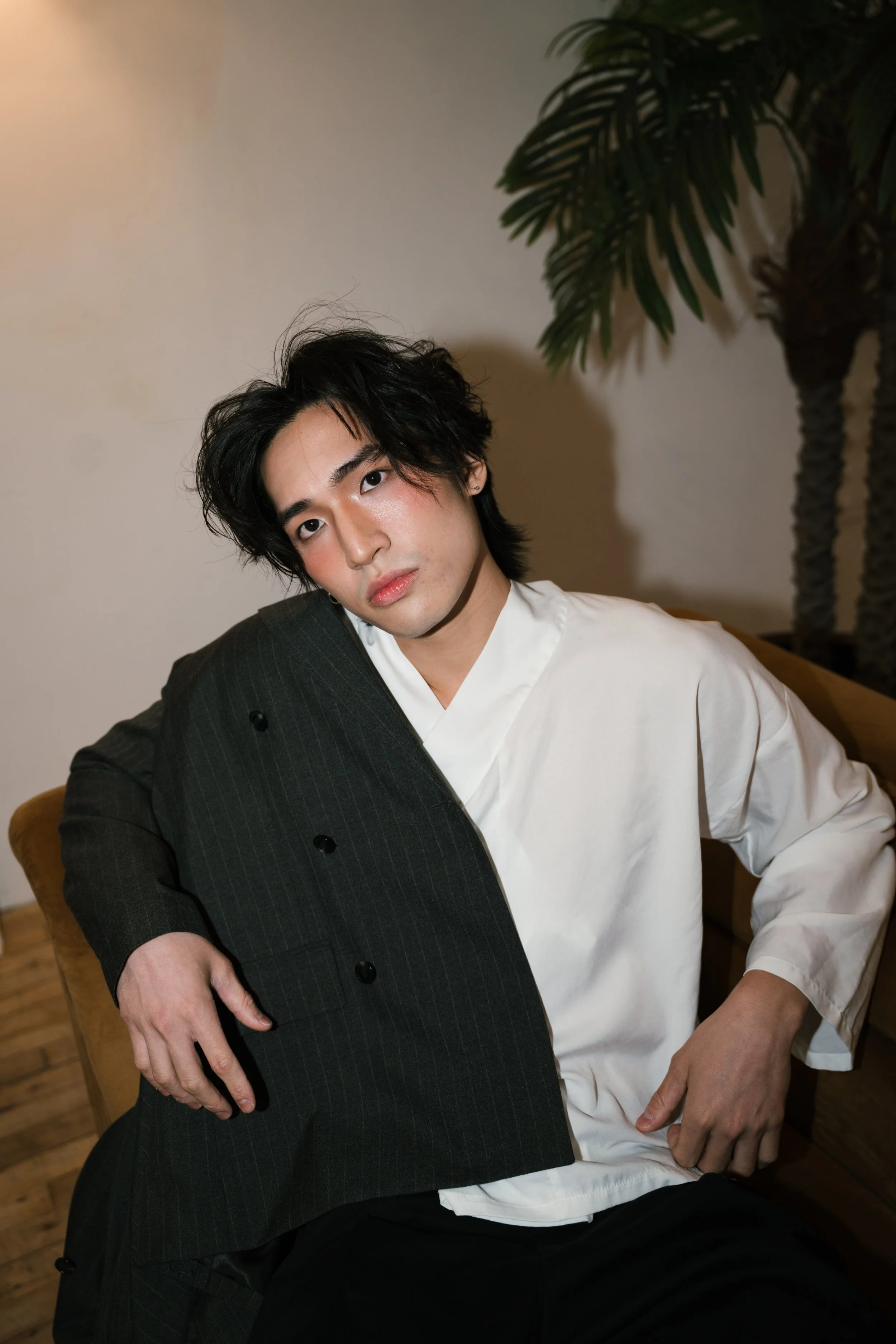 A young man with black hair, dressed in a black pinstripe suit and white shirt, sitting on a chair with a neutral background and a large green leafy plant nearby.