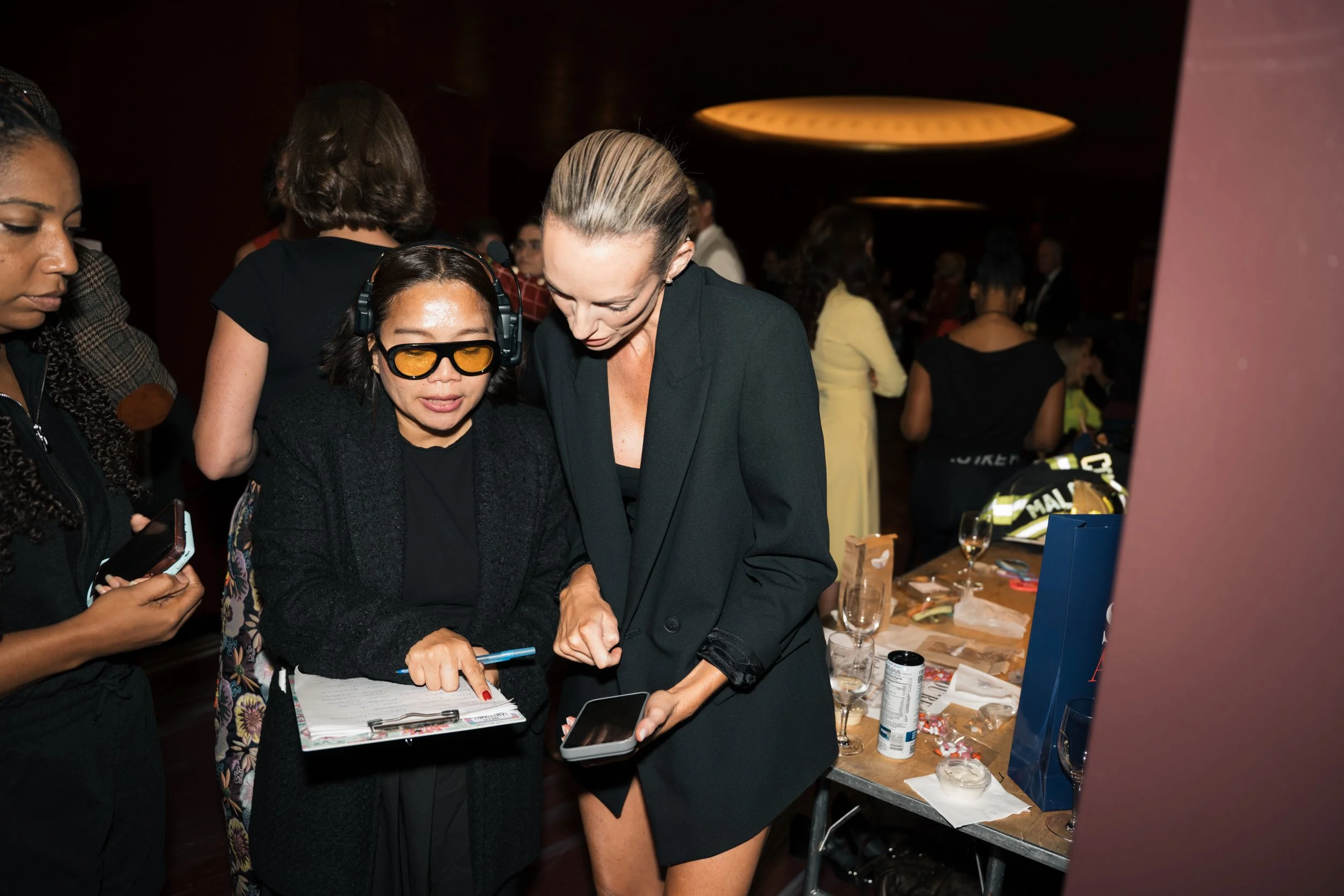 Two women engaged in conversation at a social event, with one woman wearing glasses with orange lenses and headphones, and the other holding a smartphone and pointing at a clipboard, in a dimly lit room with other guests and a table of snack items in