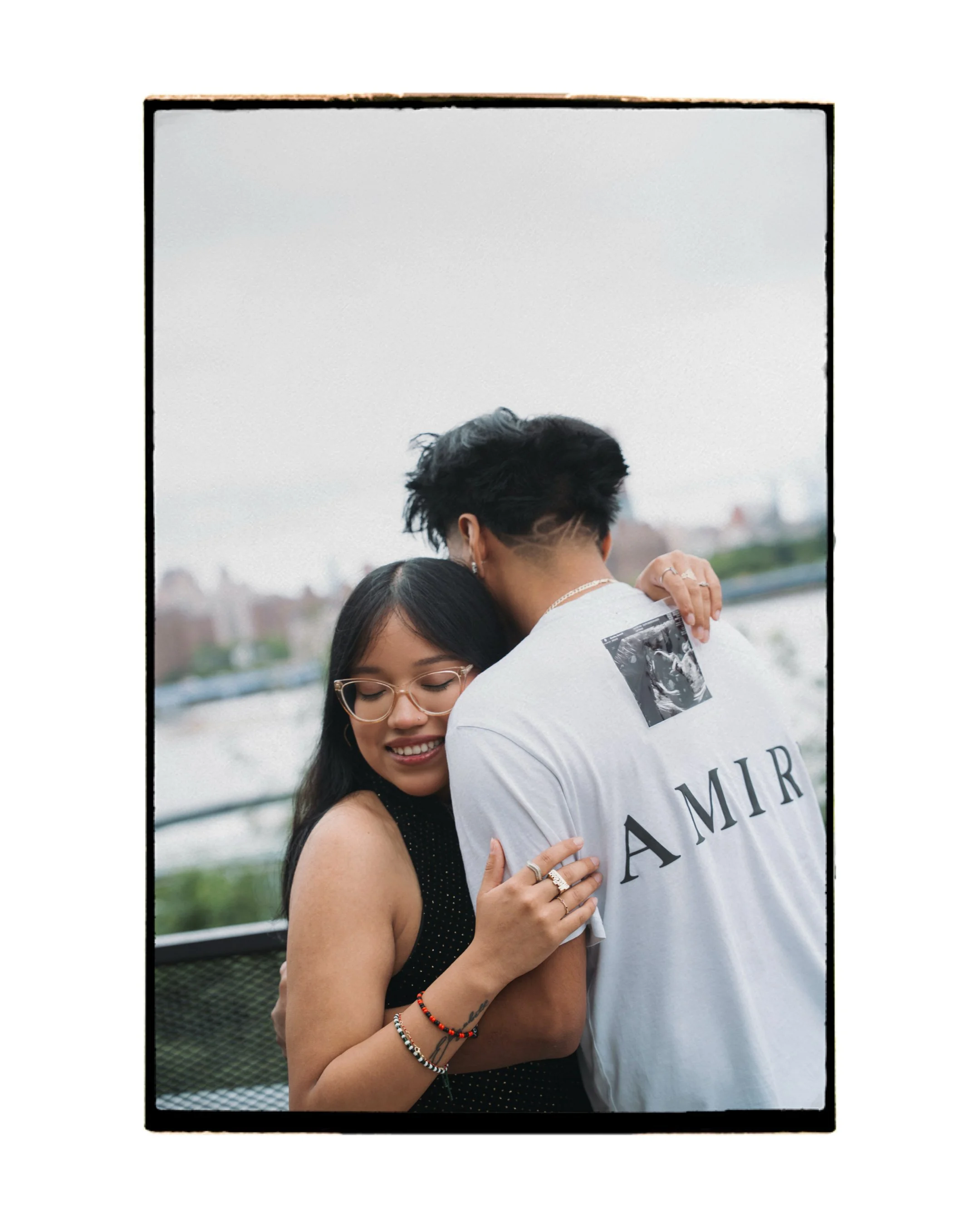 Two people hugging outdoors with a cityscape in the background. The woman has long dark hair, glasses, and multiple bracelets, and the man has short dark hair and is wearing a white t-shirt with text and an image on the back.