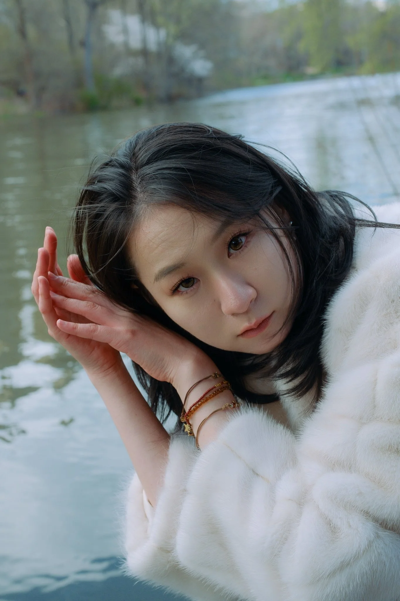 A young woman with dark hair lying on her side by a river, wearing a white furry coat, with trees in the background.