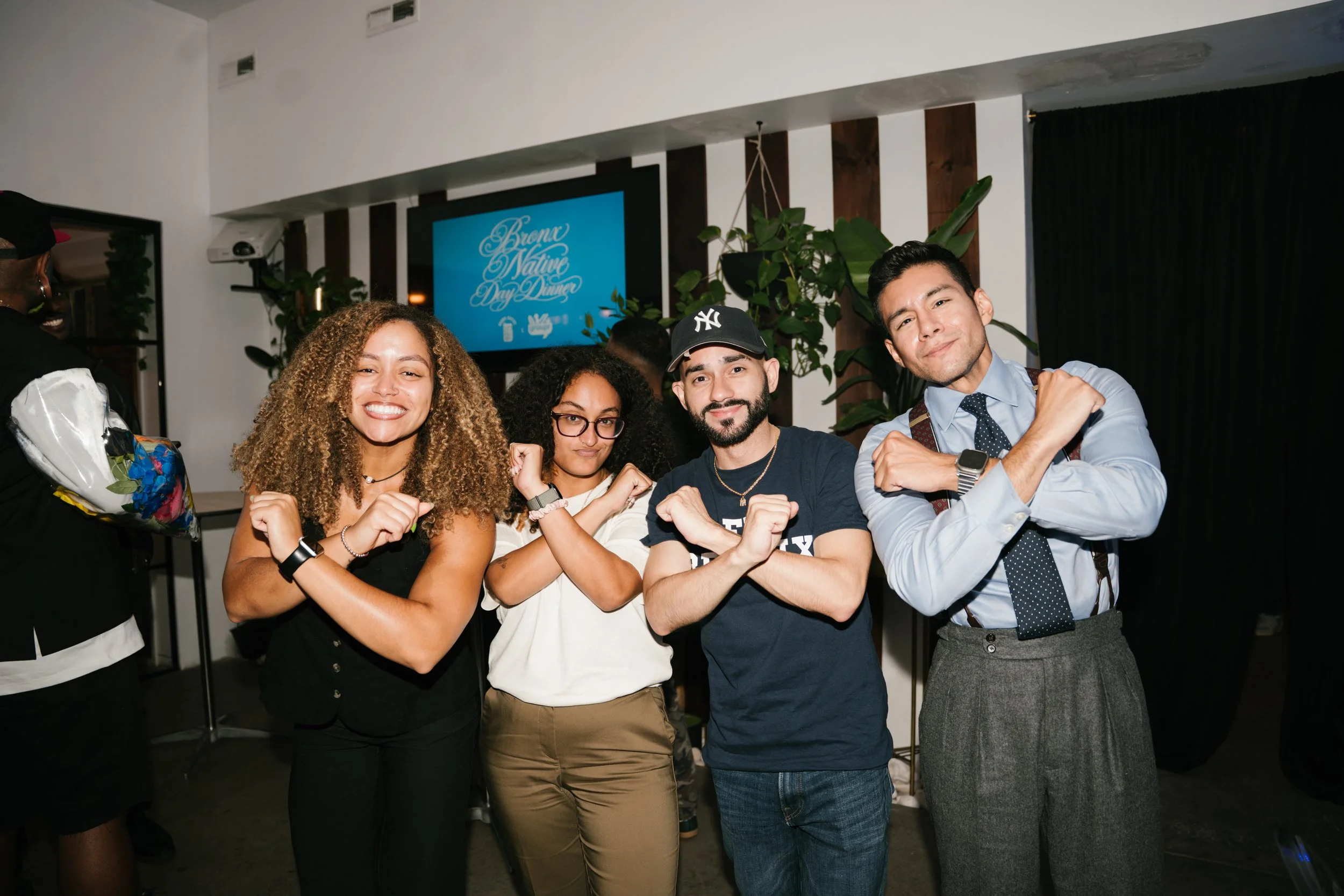 Five diverse people posing with fists clenched at a social gathering, with a digital screen in the background displaying 'Brown Native Day Dinner.'