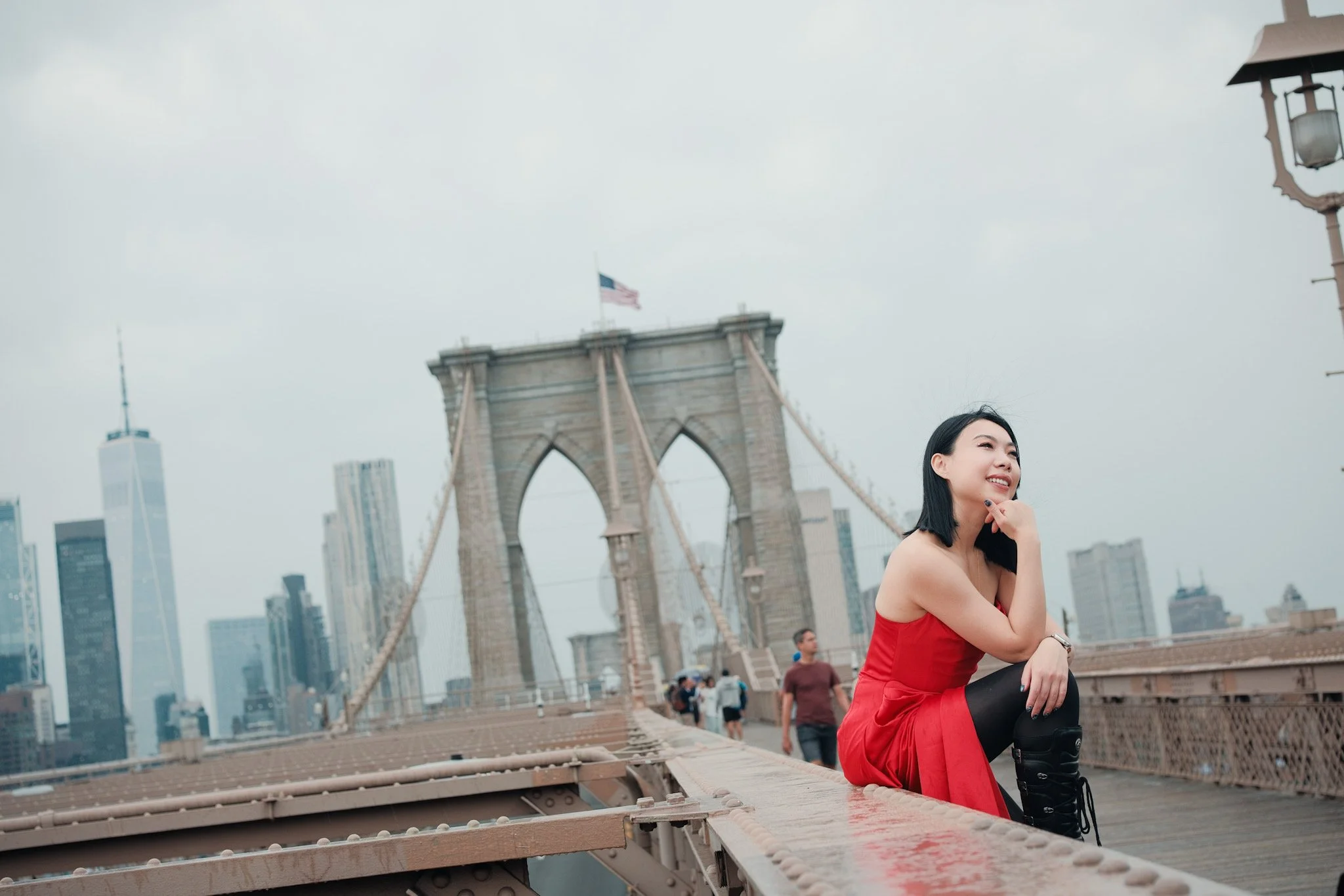 A woman in a red dress sitting on the Brooklyn Bridge with NYC skyline and the Brooklyn Bridge in the background, overcast sky.