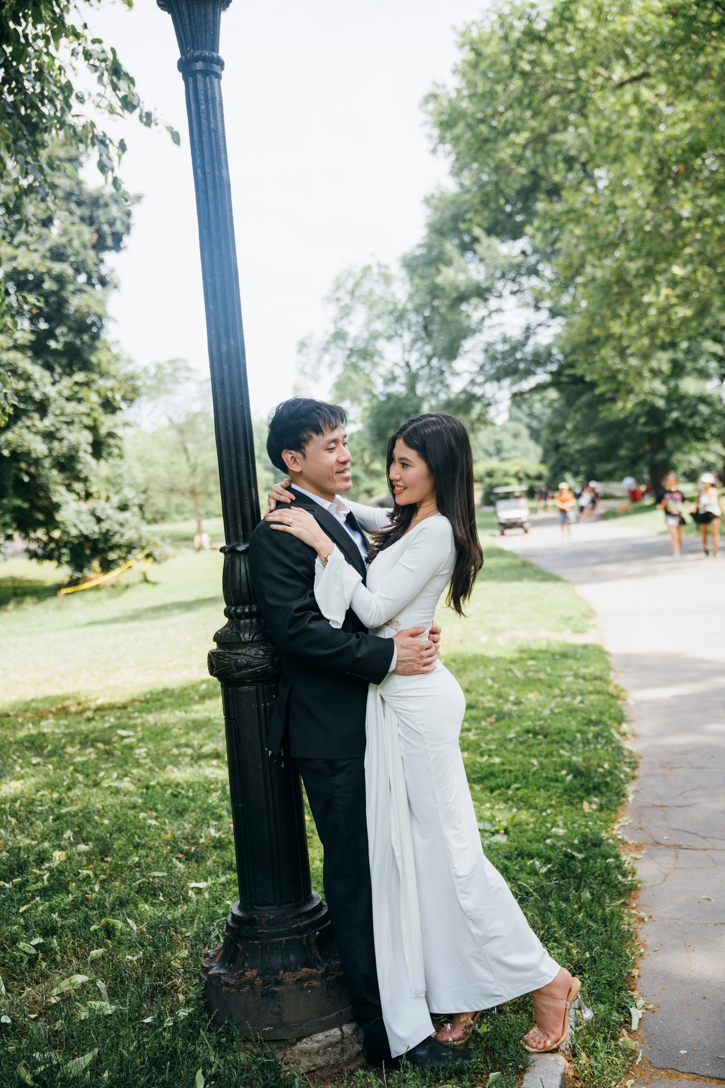 A couple dressed in formal attire, the man in a black suit and the woman in a white dress, embracing each other under a lamppost in a park with green trees and grass, and people walking in the background.