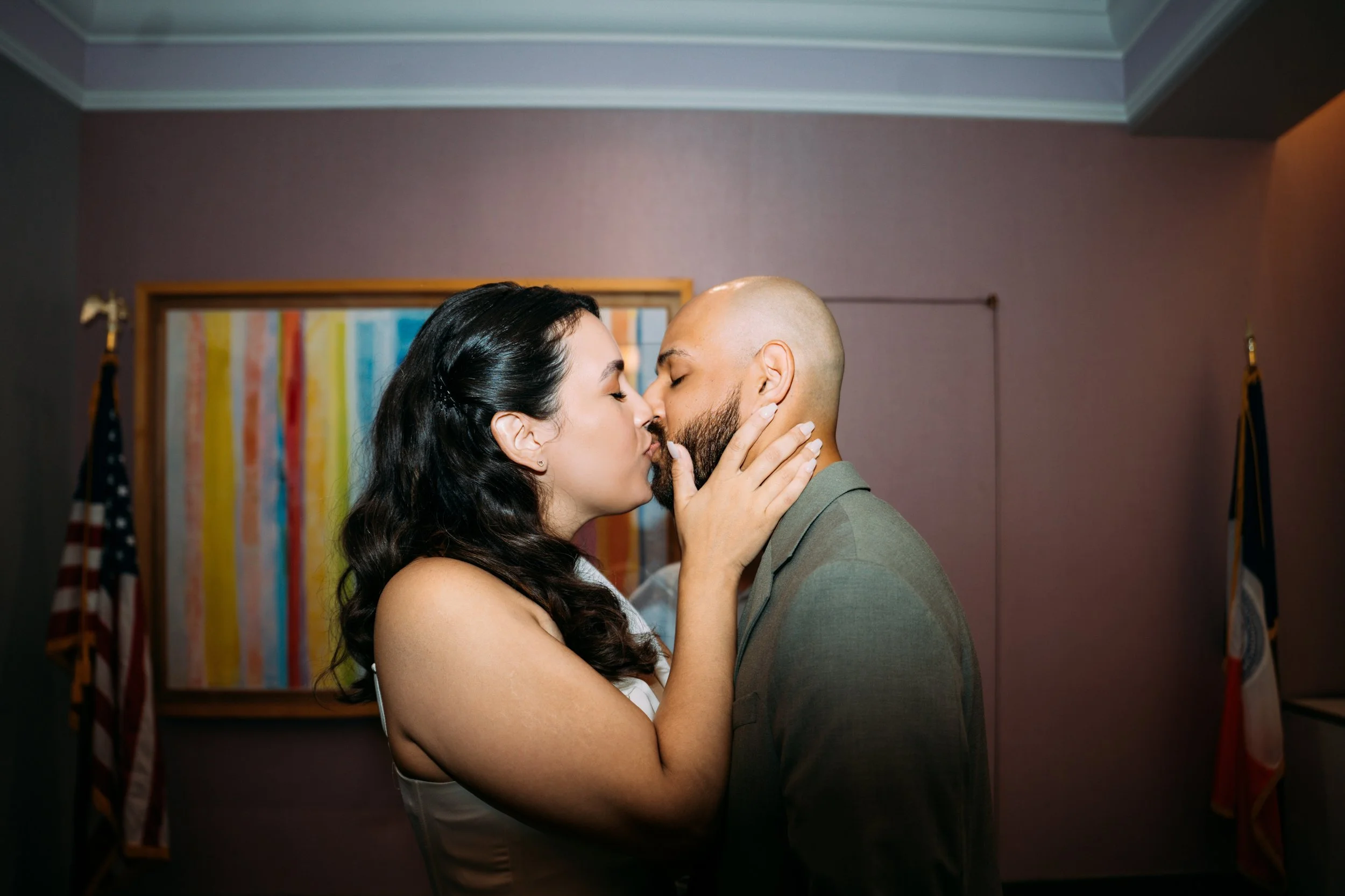 A couple shares a kiss in an indoor setting, with American flags and colorful ribbons in the background.