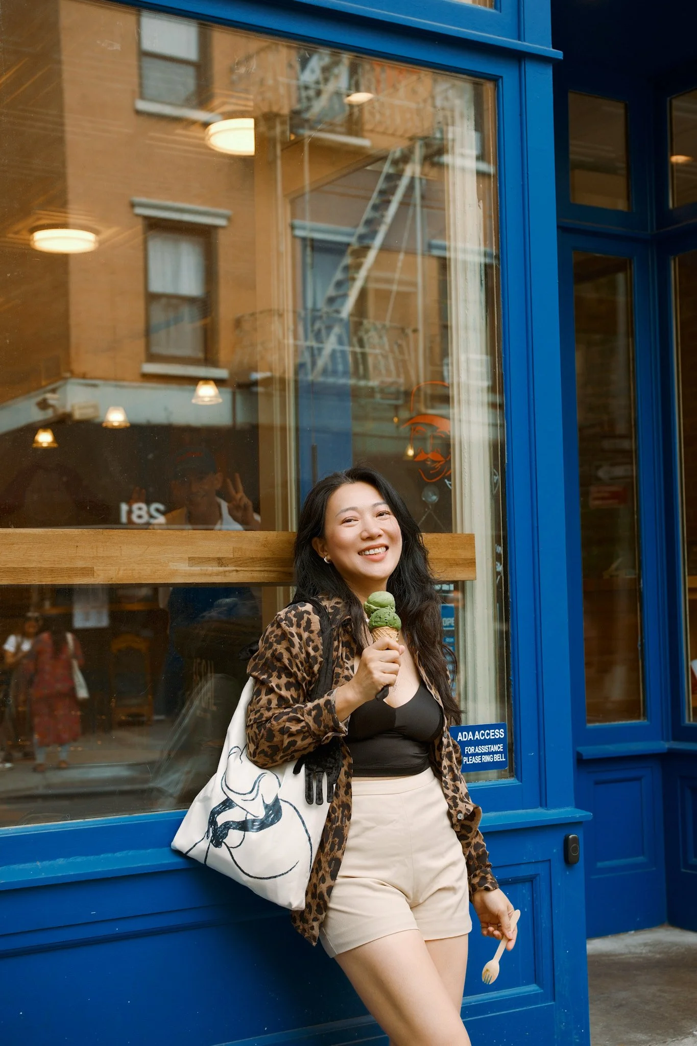 A woman with long dark hair, wearing a leopard print jacket and beige shorts, standing outside a blue storefront, holding an ice cream cone, and smiling at the camera.