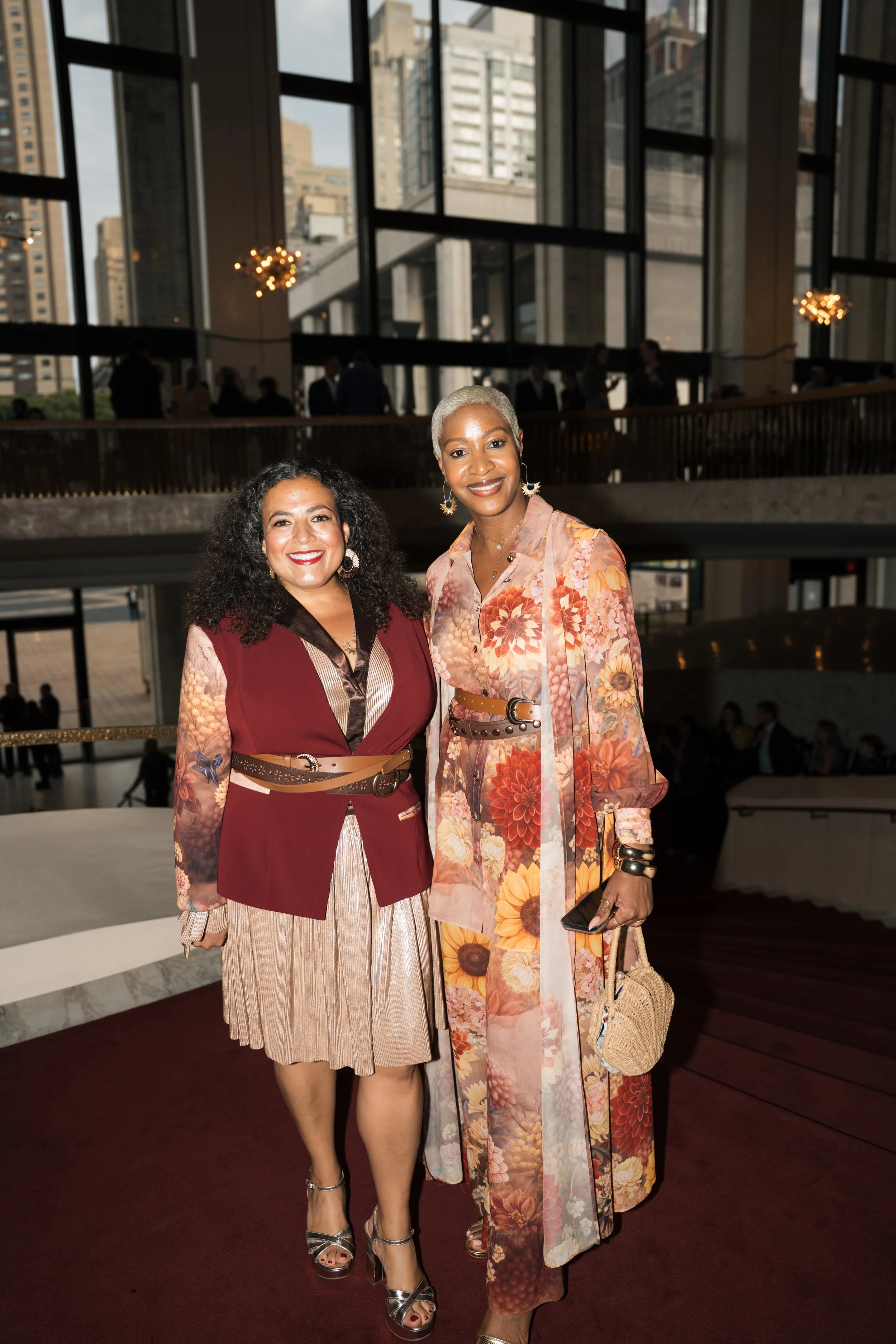 Two women stand together and smile in a modern indoor setting with large windows and cityscape views. The woman on the left has dark curly hair and is wearing a red vest over a patterned blouse and beige skirt. The woman on the right has short silver