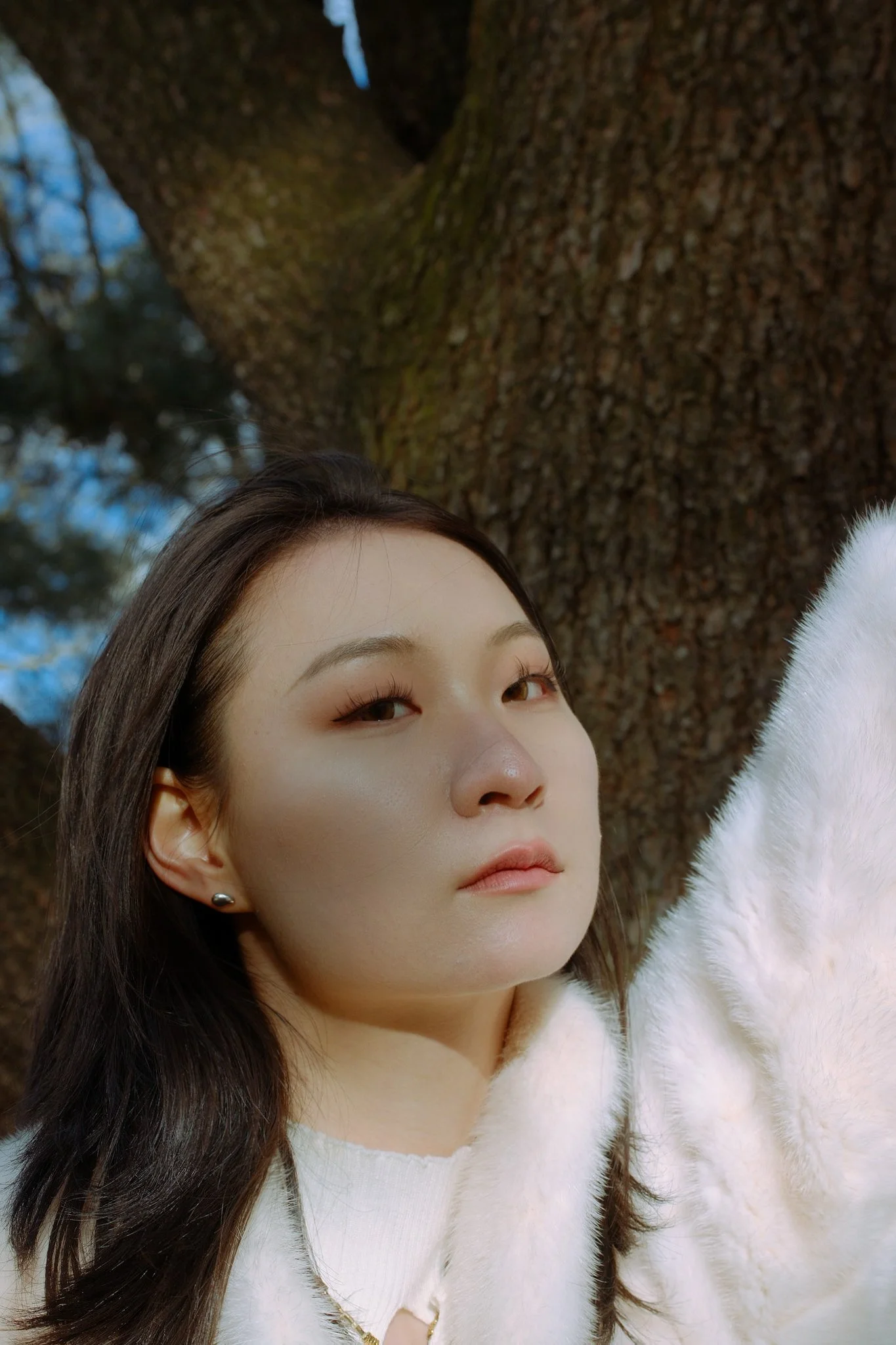 A close-up portrait of an Asian woman with long dark hair, wearing a white fuzzy coat, standing outdoors in front of a large tree with textured bark, and looking slightly to her right.